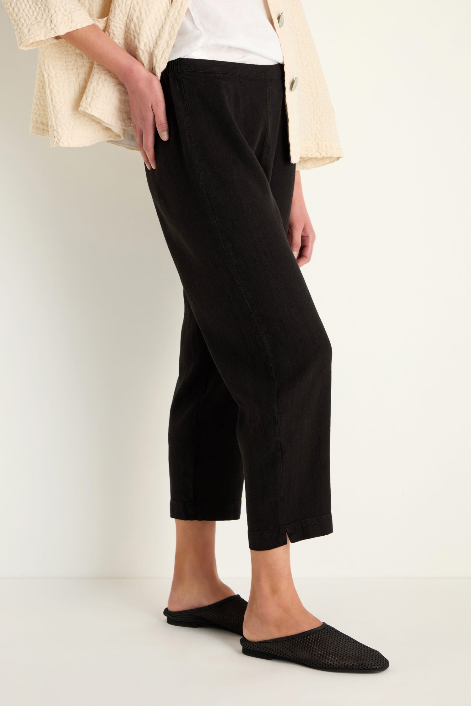A person stands against a white background wearing Twisted Linen Slim Trousers in black, a white top, a cream textured jacket, and black flat shoes.