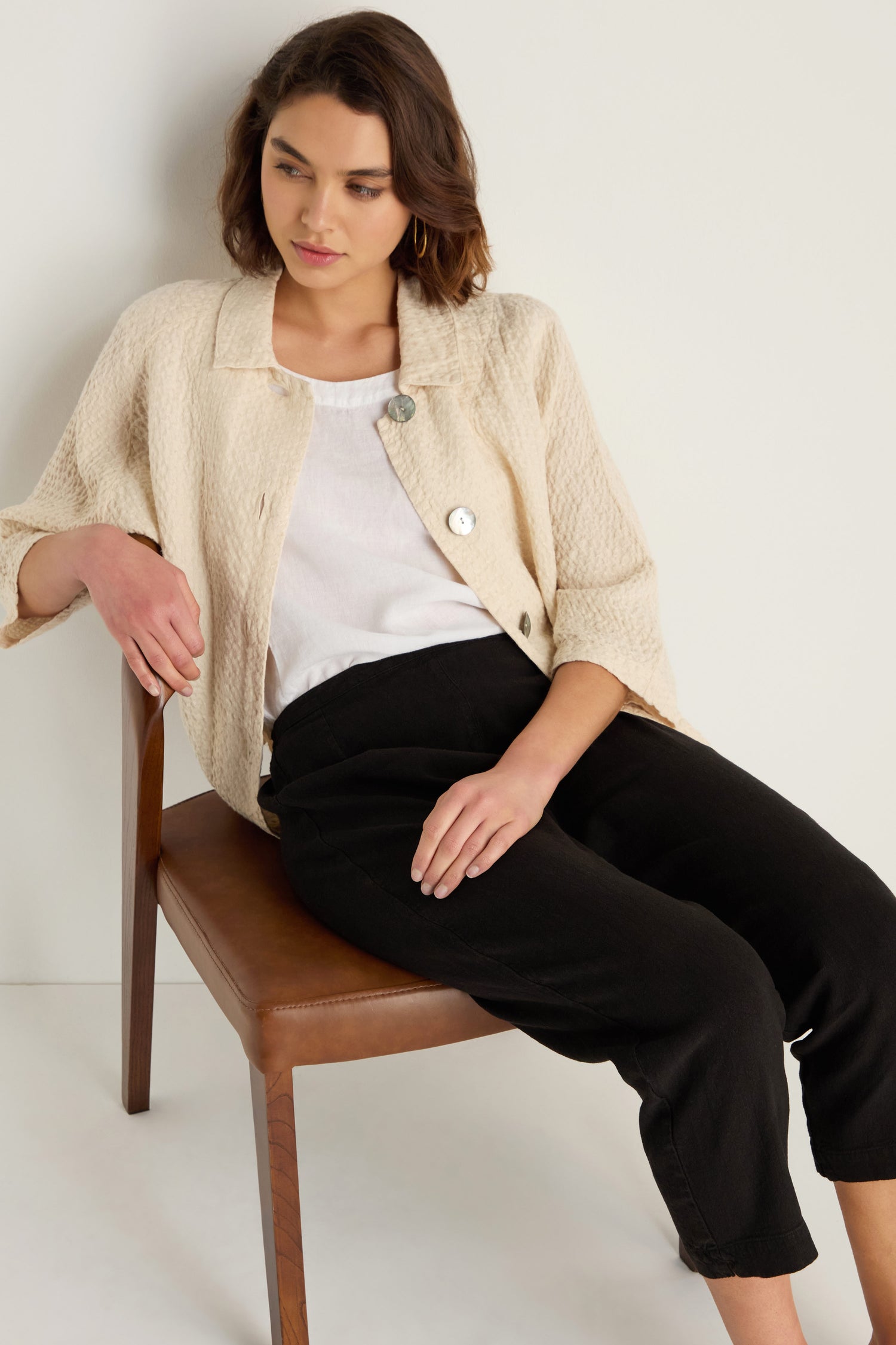 A woman sits on a brown chair against a white wall, wearing a cream textured jacket, white shirt, and Twisted Linen Slim Trouser.
