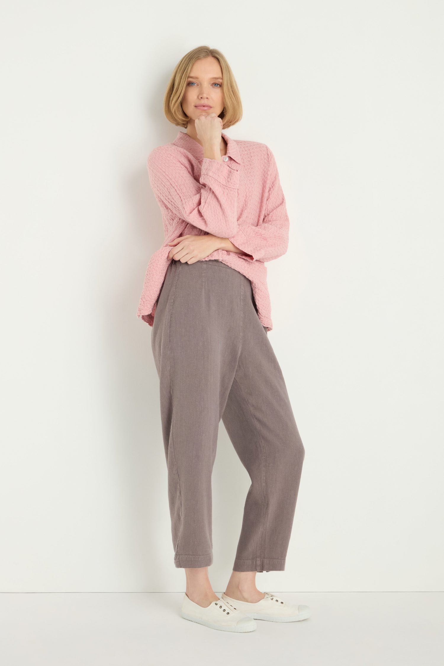 A person stands against a white wall, wearing a pink textured long-sleeve top, Twisted Linen Slim Trouser in gray, and white sneakers, with one hand resting on their chin.