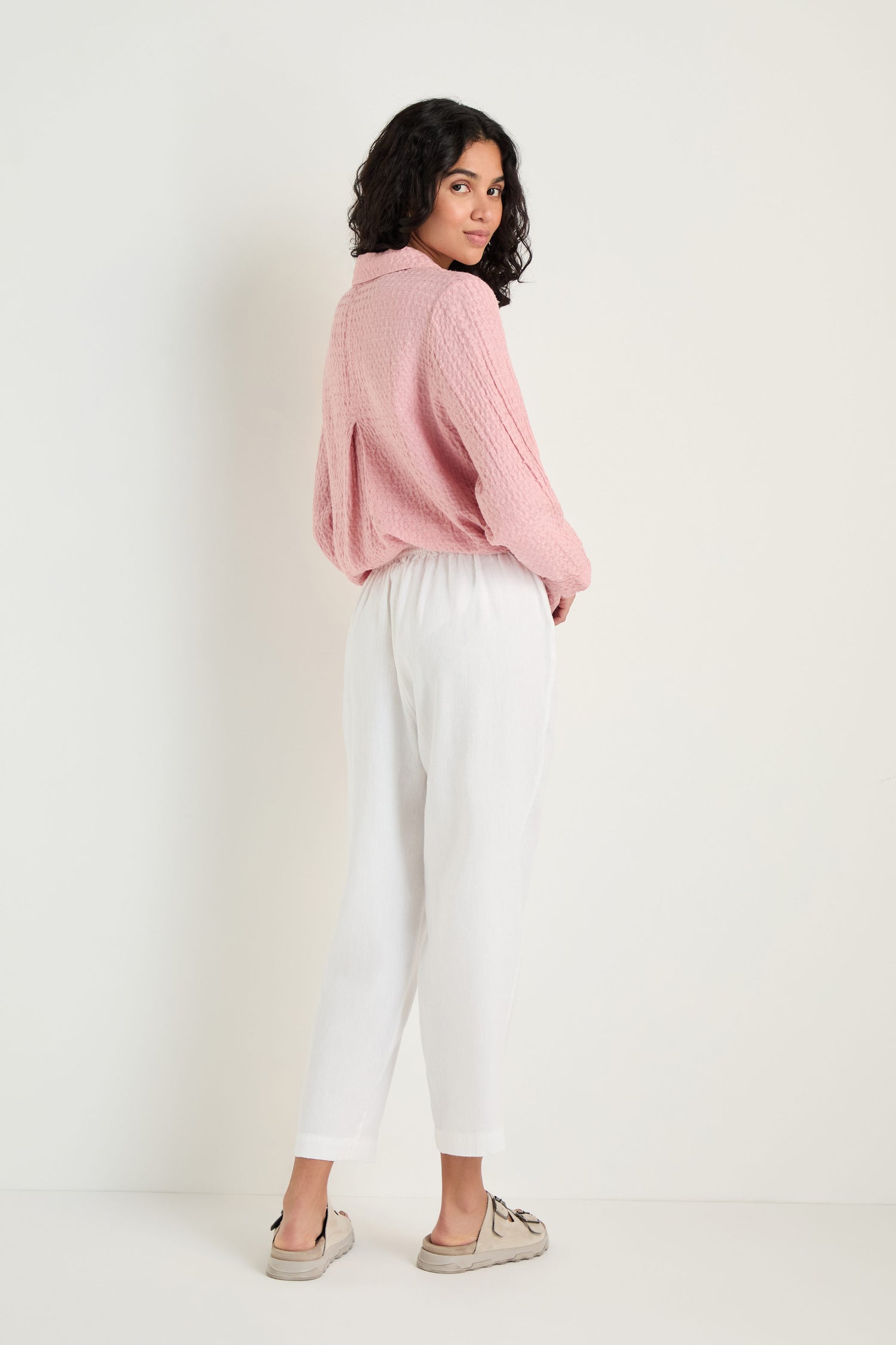 A person stands against a plain light background, wearing a pink textured sweater and Twisted Linen Slim White Trouser in an ankle-grazing length with beige loafers, looking back over their shoulder.