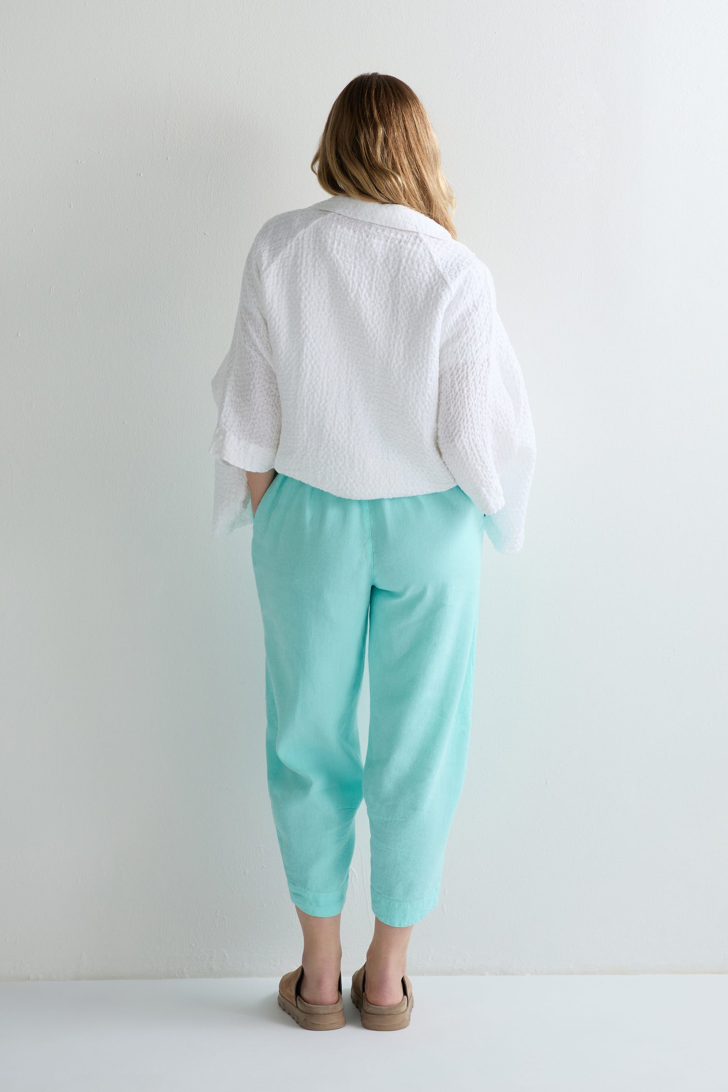 A person stands with their back to the camera, wearing a white textured top, brown sandals, and Mint Twisted Linen Crop Bubble Trousers featuring breathable twisted linen fabric and a bubble shape, against a plain light-colored wall.