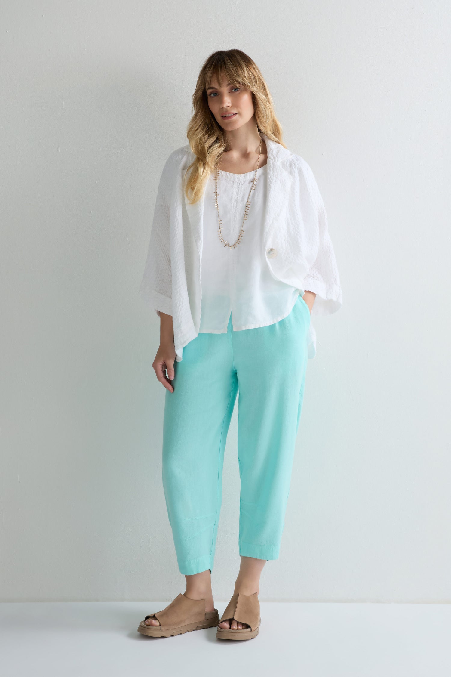 A woman stands against a plain white wall wearing a white blouse, loose white jacket, Mint Twisted Linen Crop Bubble Trouser, beige sandals, and a long necklace. The bubble-shaped trouser is crafted from breathable twisted linen.