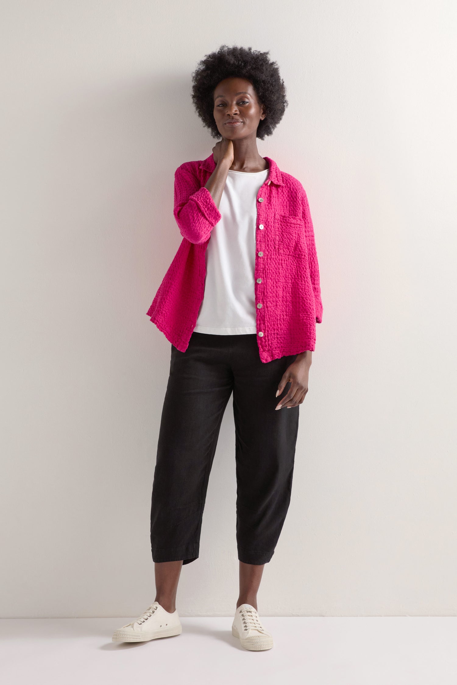In front of a white wall, a person poses casually in layered attire with a pink shirt over a white top, complemented by black Twisted Linen Crop Bubble Trousers and clean white sneakers.