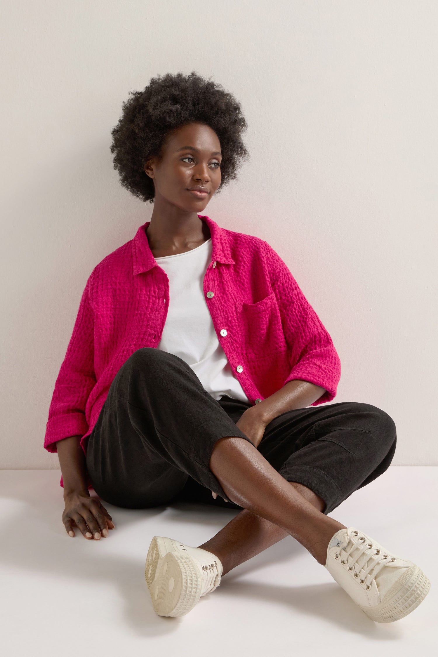 A person with curly hair casually sits on the floor in a bright pink jacket, white shirt, and Twisted Linen Crop Bubble Trouser. They gaze off to the side with a neutral expression, highlighting the outfit's versatile layering potential.