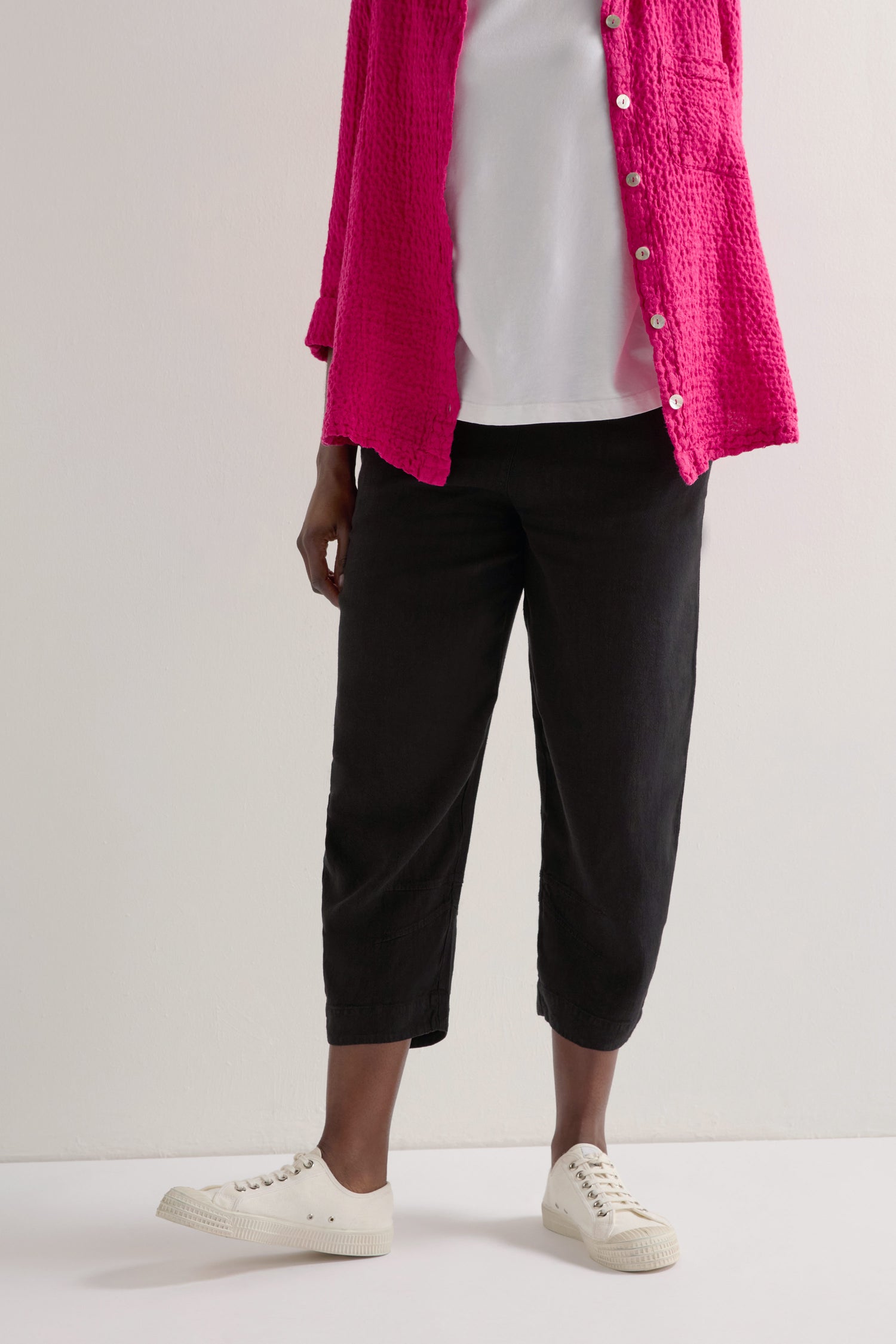 A person in a pink textured shirt, white T-shirt, and black Twisted Linen Crop Bubble Trouser paired with white sneakers creates a relaxed look against a light background.
