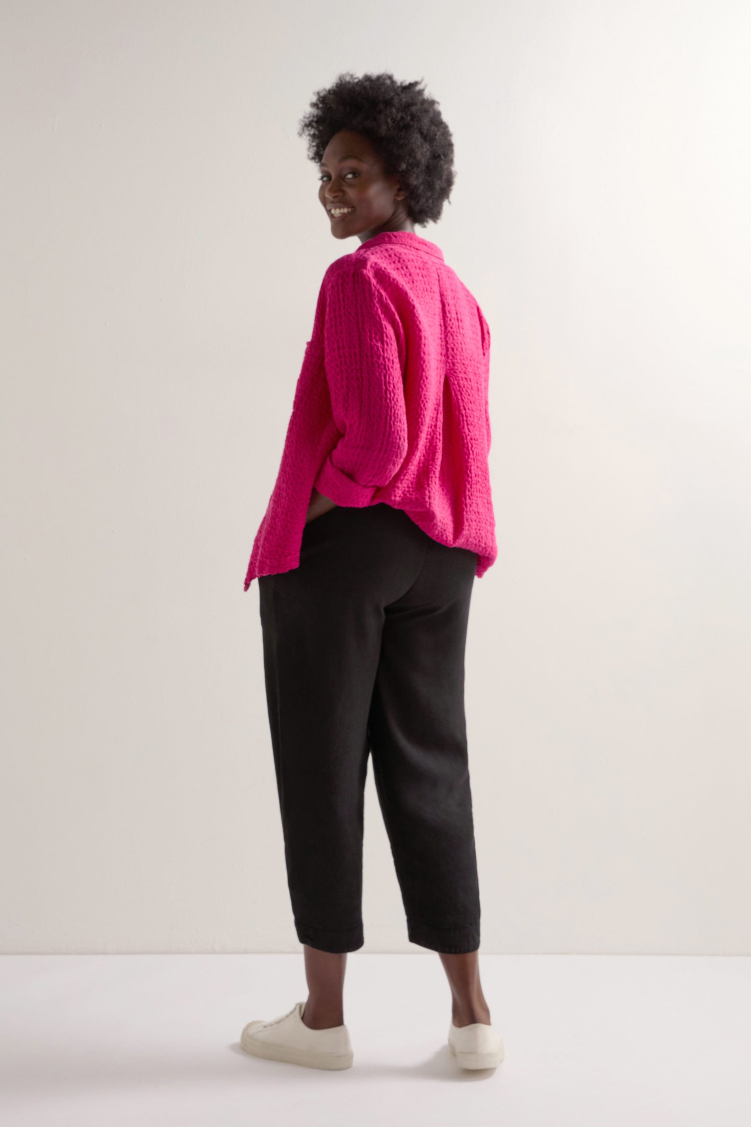 Wearing a bright pink sweater and Twisted Linen Crop Bubble Trousers with white shoes, a person presents a relaxed look against a plain background.