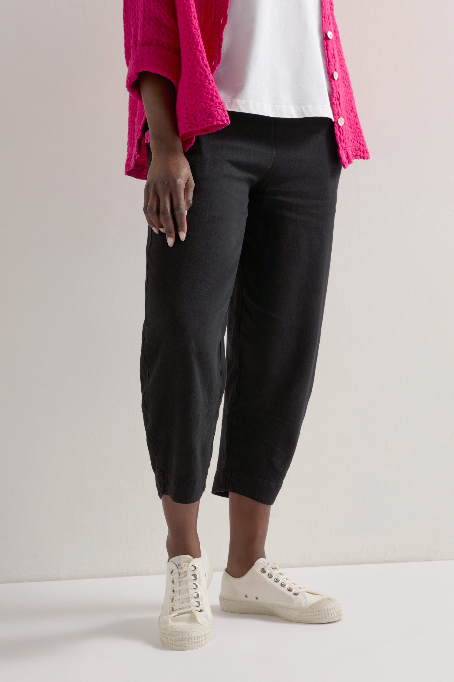 Wearing Twisted Linen Crop Bubble Trousers, a crisp white shirt, a pink sweater, and white sneakers, the person displays a versatile layering style against a plain background.