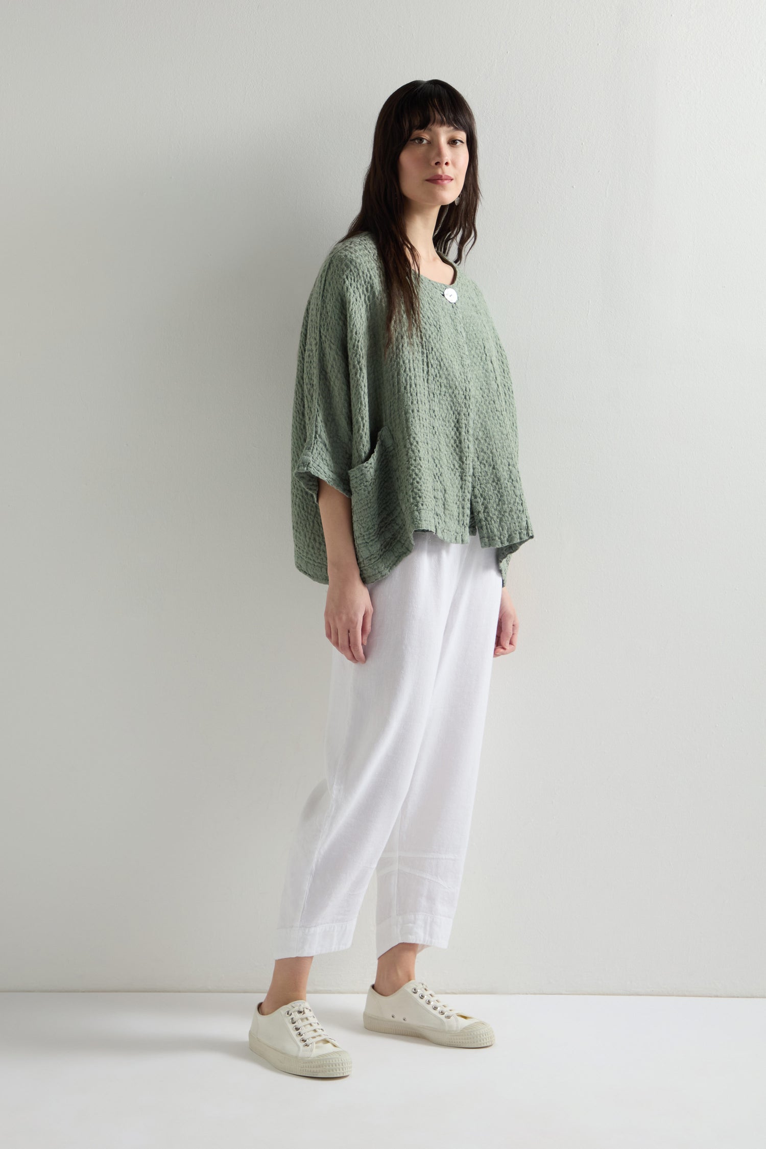 A person stands against a plain background, wearing a green textured top and Twisted Linen Crop Bubble Trousers with an elasticated waistband, paired with white sneakers.