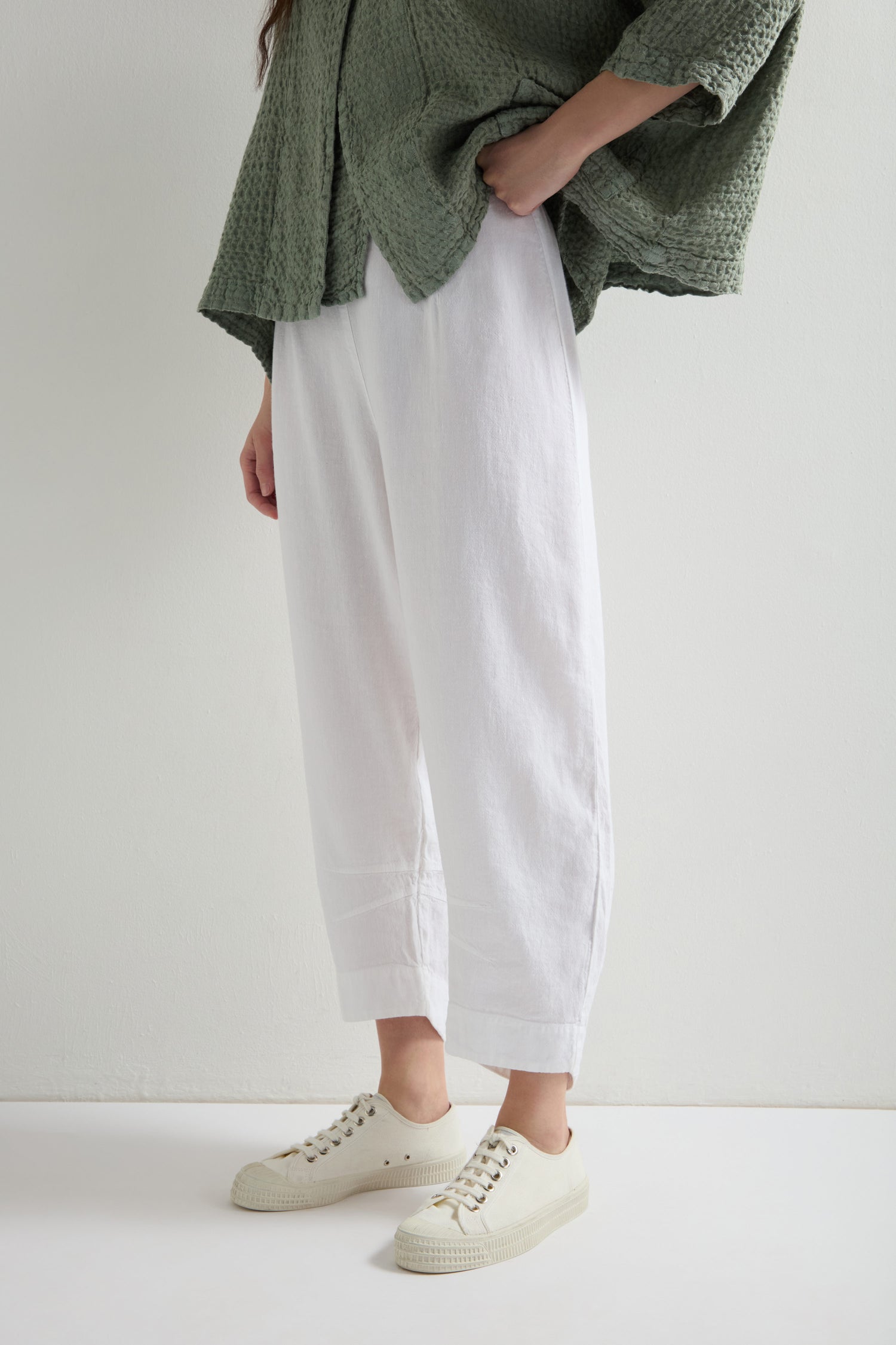 A person in a green textured top and cream-colored sneakers stands against a plain light background. Their white Twisted Linen Crop Bubble Trousers with an elasticated waistband create a relaxed yet stylish vibe.