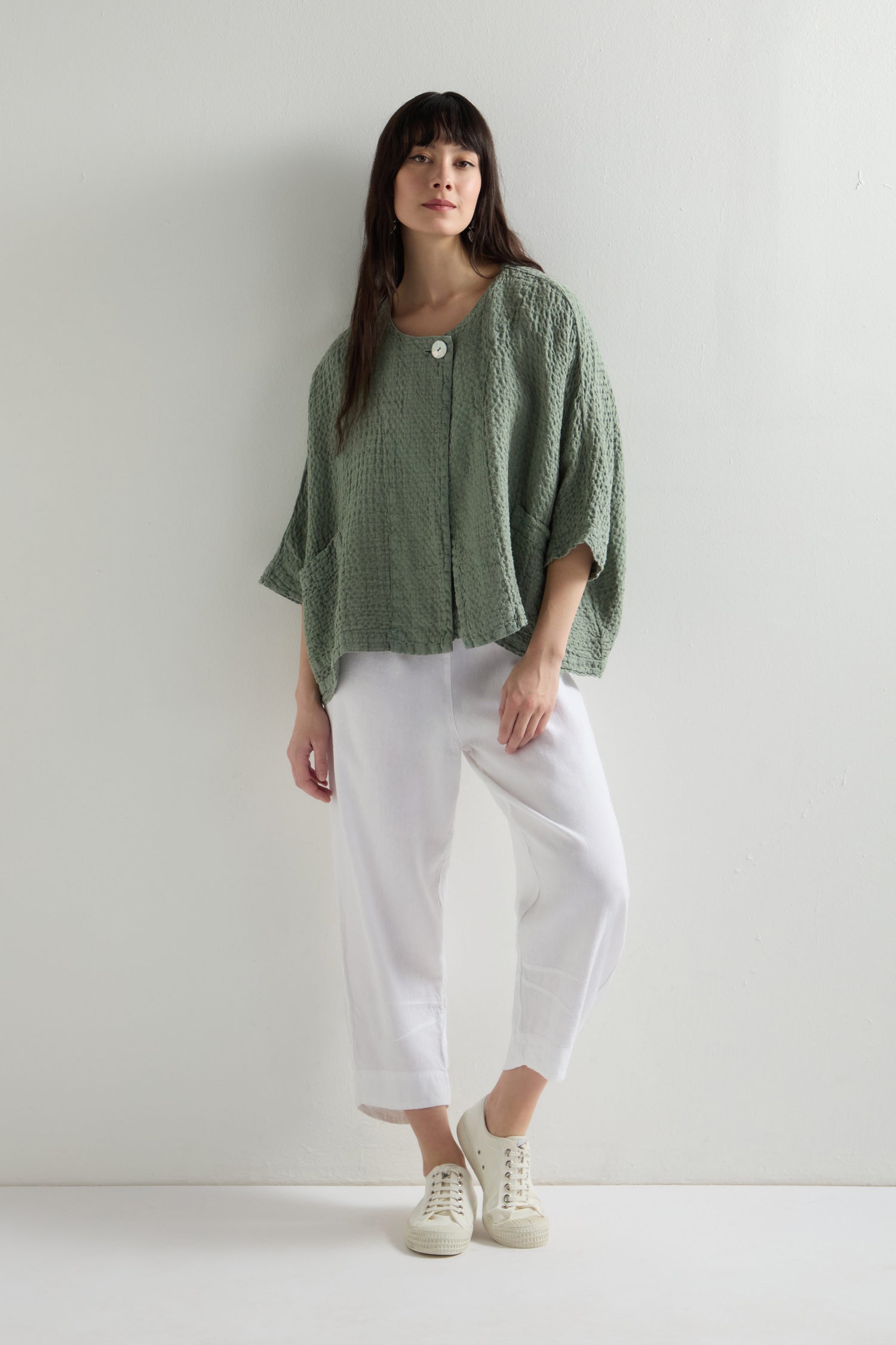 A person stands against a white wall wearing an elegant green textured top paired with Twisted Linen Crop Bubble Trouser, featuring an elasticated waistband, and chic white sneakers.