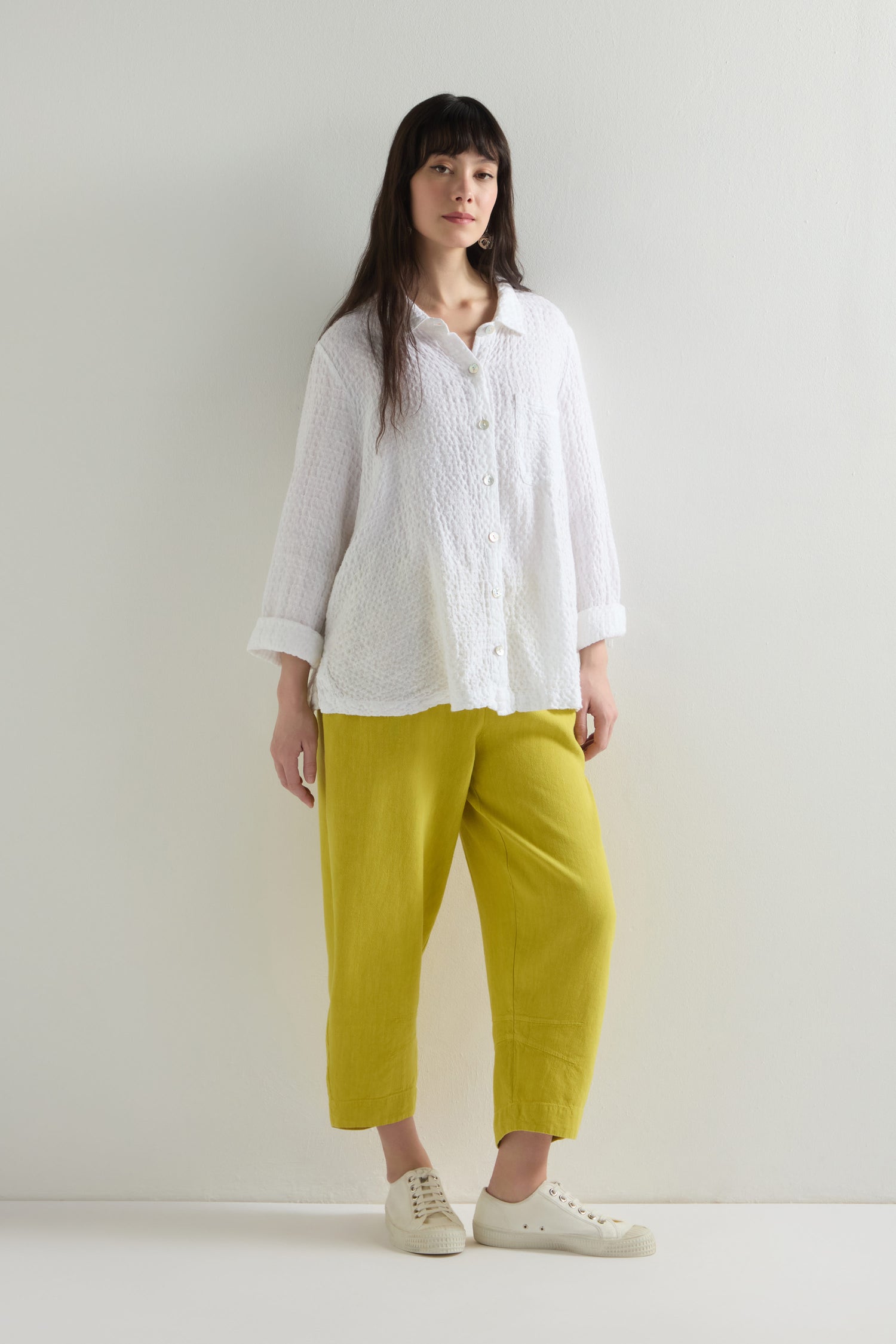 A person stands against a white wall, looking relaxed in a loose white button-up shirt and Twisted Linen Crop Bubble Trouser, complemented perfectly by fresh white sneakers.
