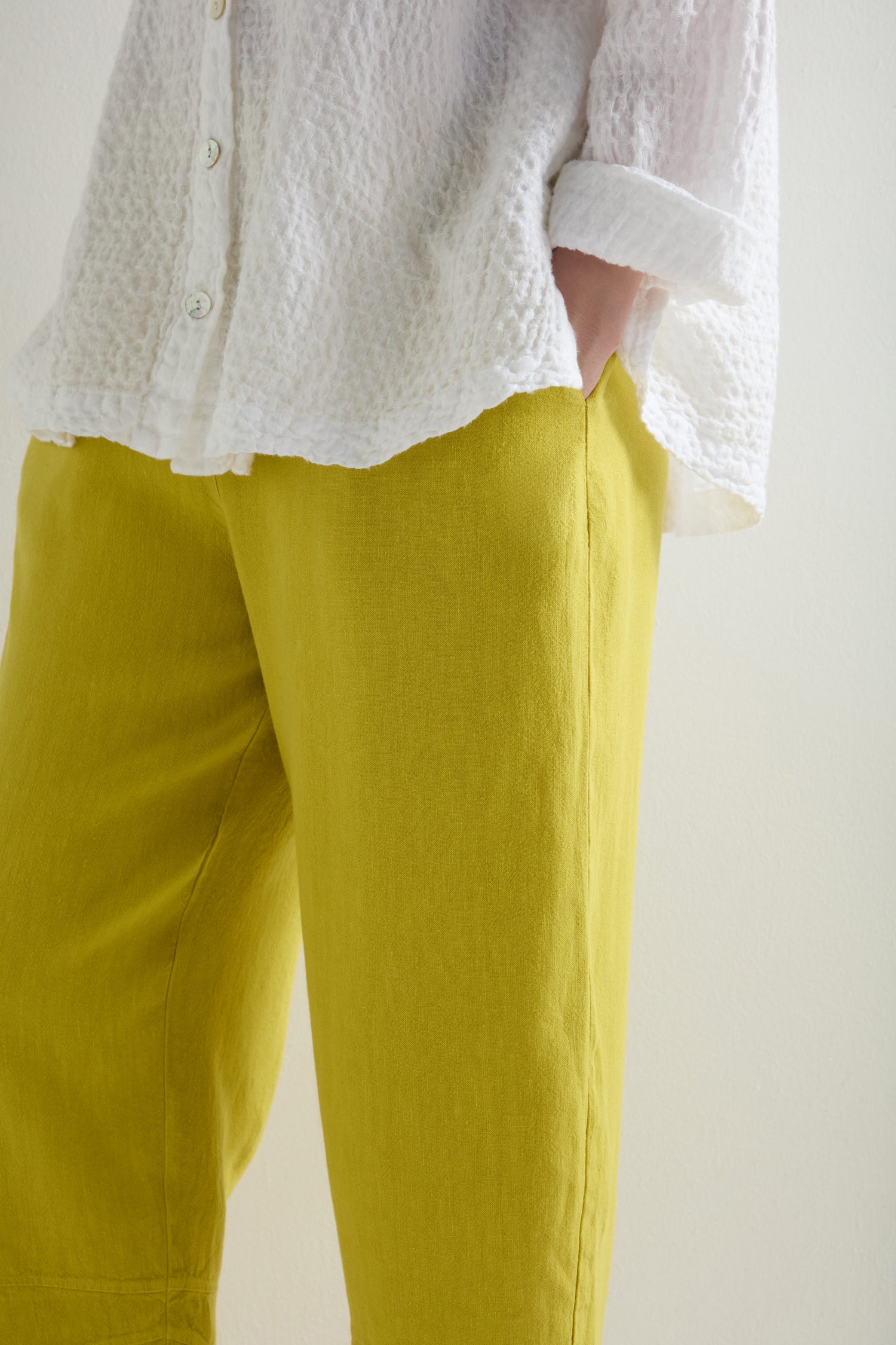 A person exudes a relaxed look, donning a textured white shirt with lively yellow Twisted Linen Crop Bubble Trousers, one hand casually resting in a pocket.