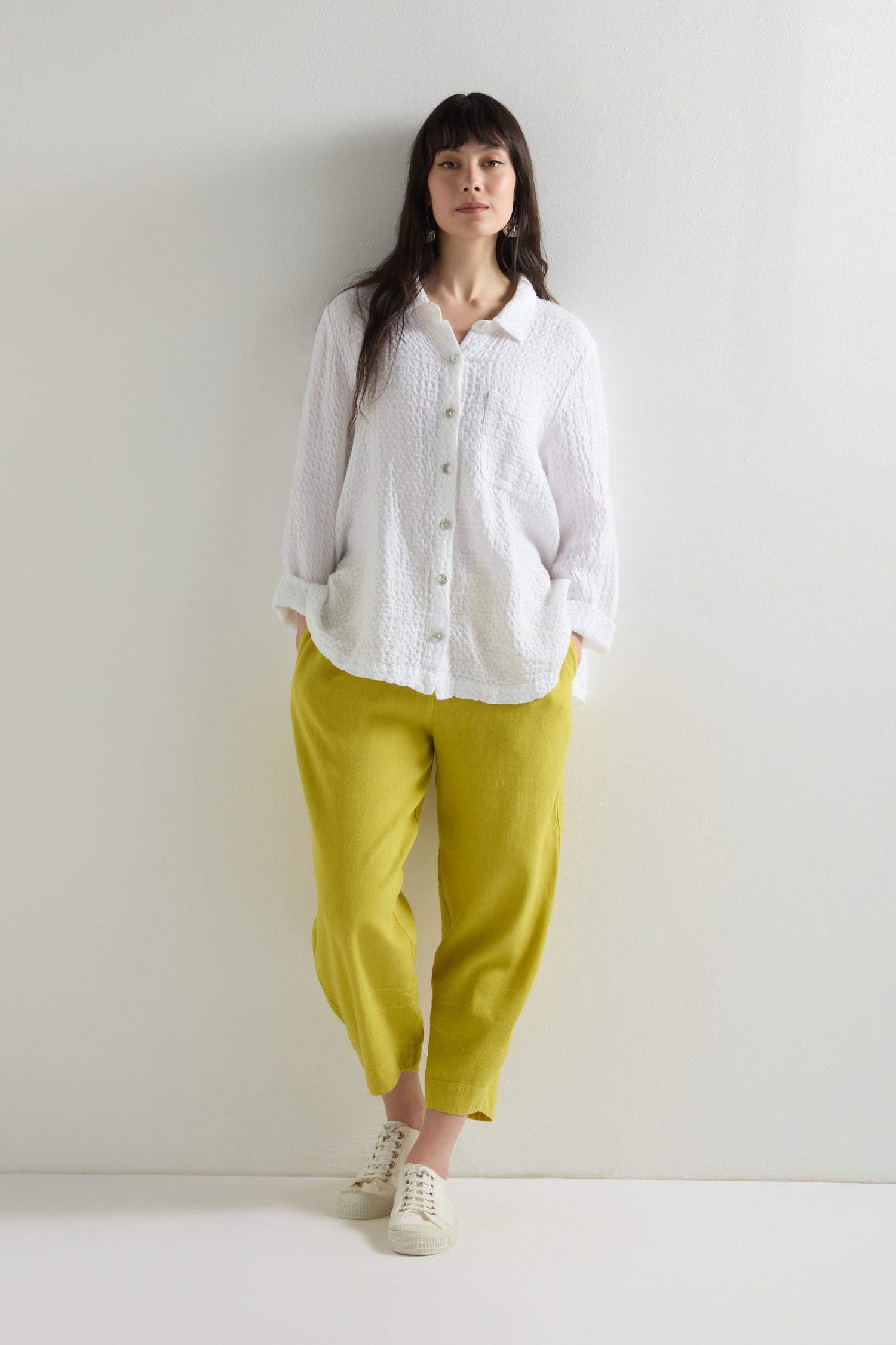 A person is casually standing with hands in pockets, wearing a white button-up shirt paired with yellow Twisted Linen Crop Bubble Trousers, which add a contemporary touch to the relaxed look against the plain background.