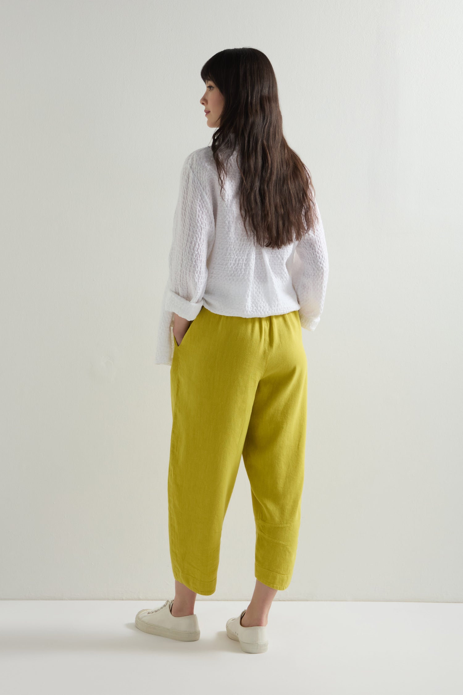 A person with long dark hair, wearing a white textured sweater and Twisted Linen Crop Bubble Trousers with an elasticated waistband, stands away against a plain white background. The outfit is completed with yellow pants and sleek white sneakers.