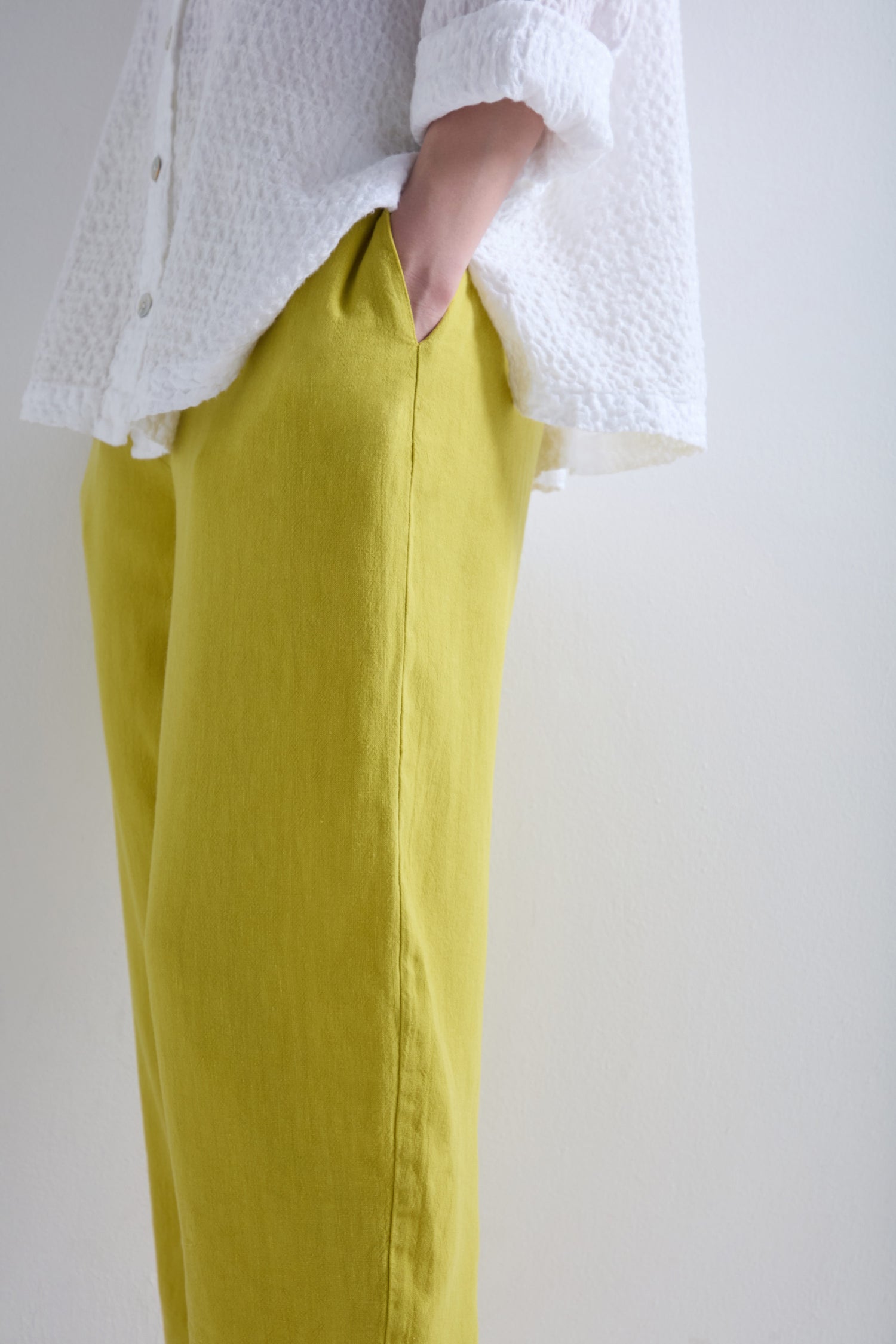Donning a white textured shirt and yellow Twisted Linen Crop Bubble Trousers, with one hand casually in the pocket, the person exudes a relaxed vibe against a plain backdrop.