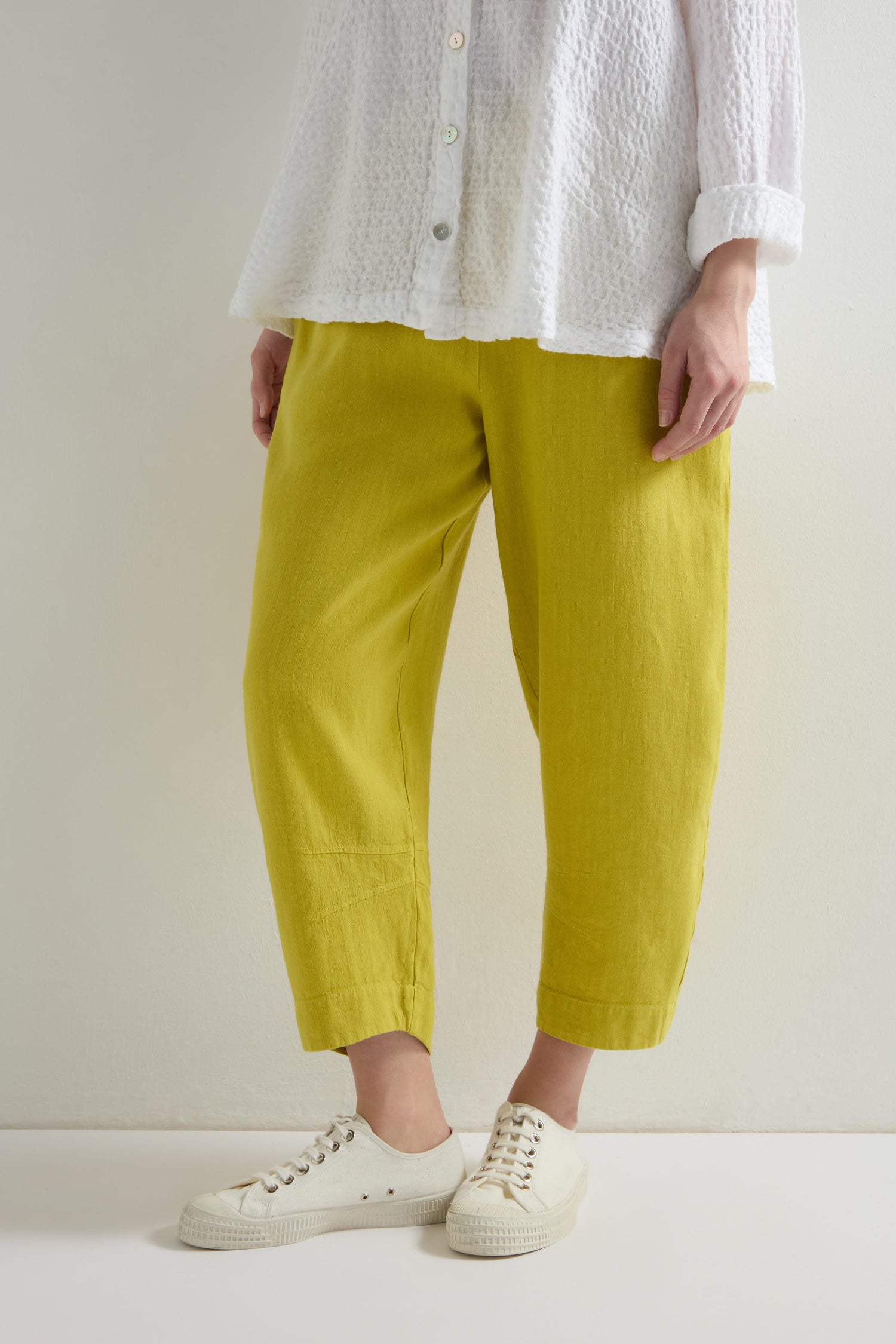 Wearing a white textured shirt and Twisted Linen Crop Bubble Trouser in bright yellow with an elastic waistband, paired with white sneakers, a person stands against a neutral backdrop.