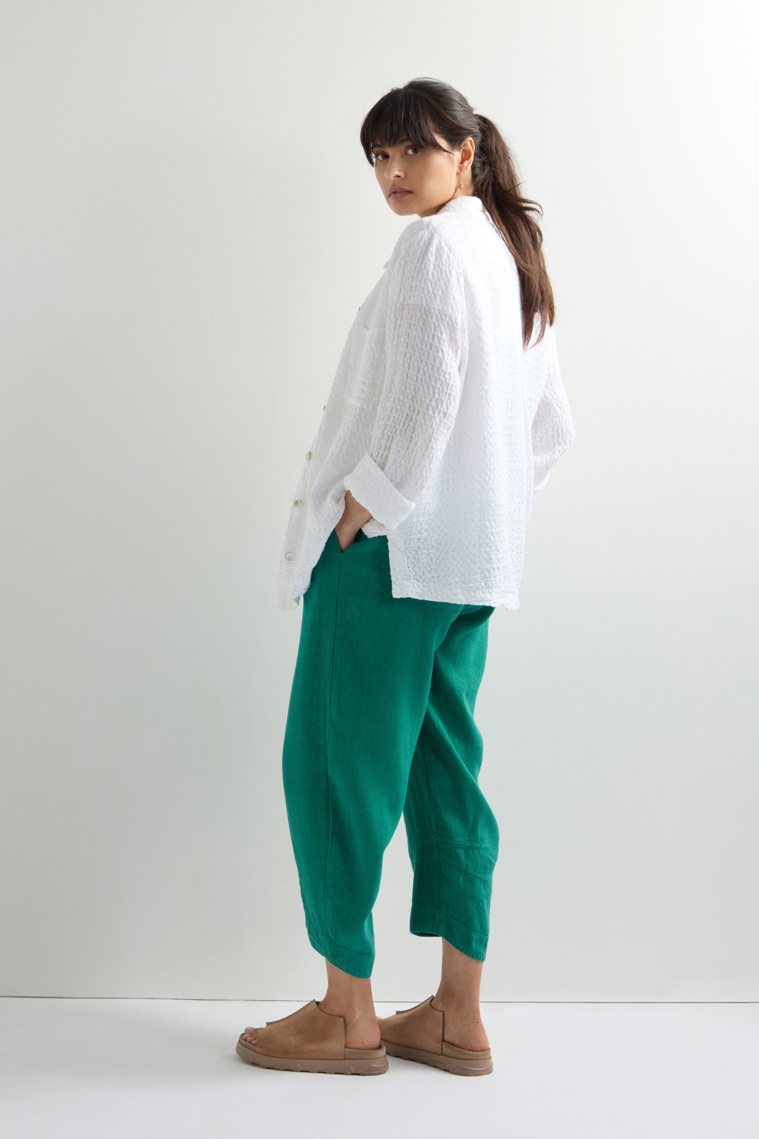 With effortless style, a person stands against a plain background wearing a white blouse, brown sandals, and green Twisted Linen Crop Bubble Trousers with an elasticated waistband. They look over their shoulder in comfort.