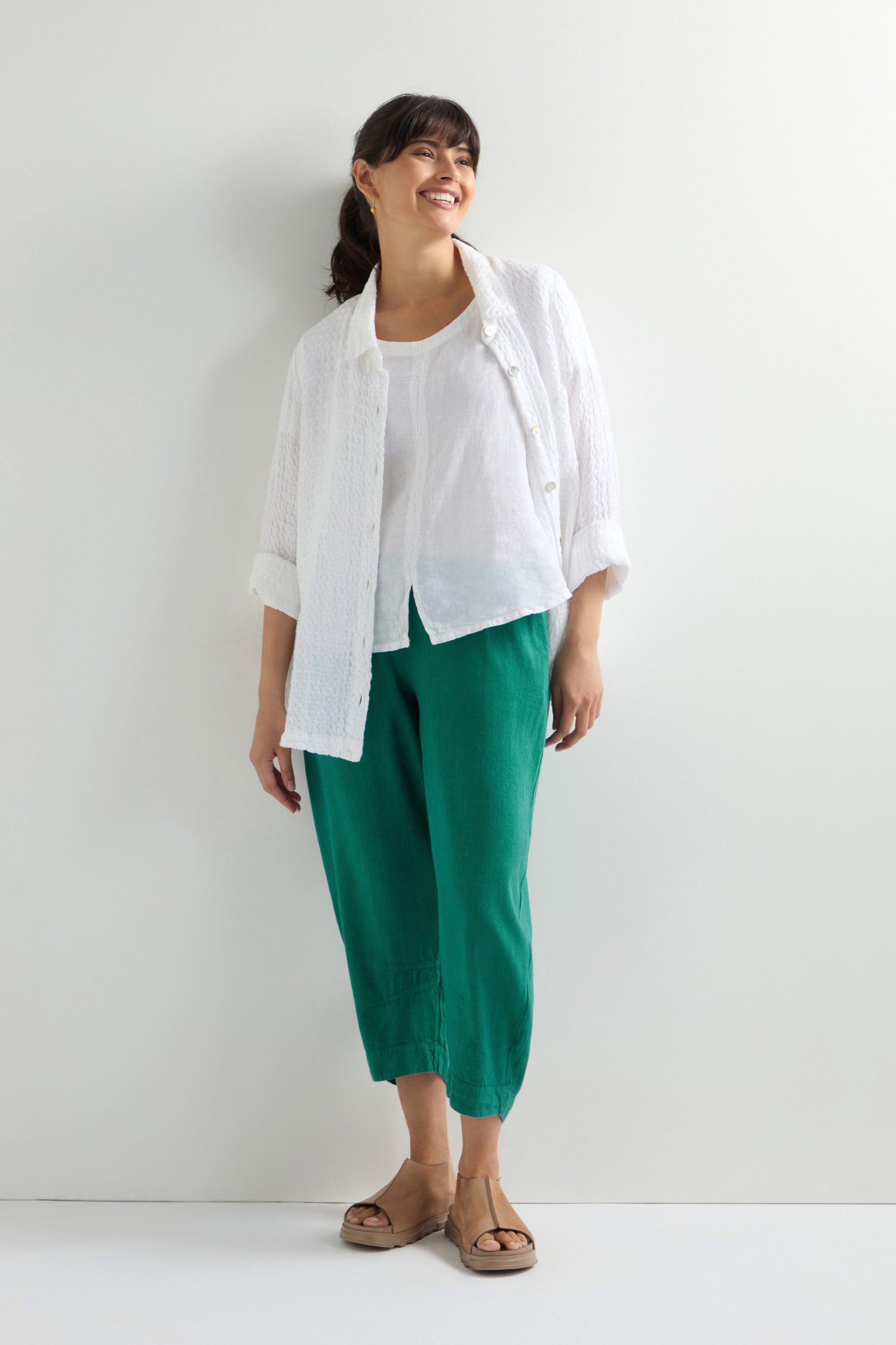 A person stands against a white wall, wearing a white shirt over a blouse and green Twisted Linen Crop Bubble Trousers paired with sandals.