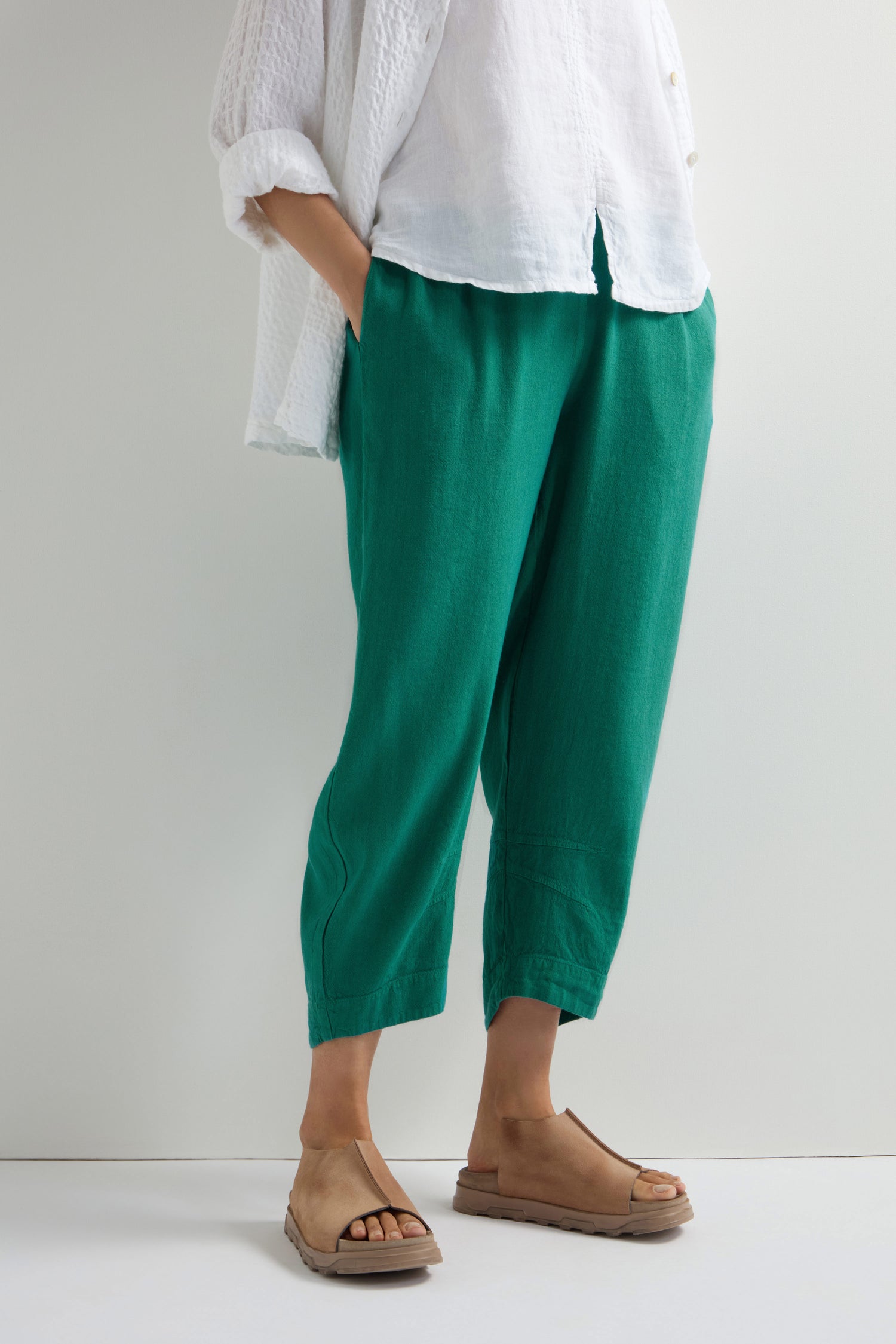 Someone is wearing a white shirt paired with Twisted Linen Crop Bubble Trousers, which have an elastic waistband, and tan sandals, focusing on their lower half against a plain background.