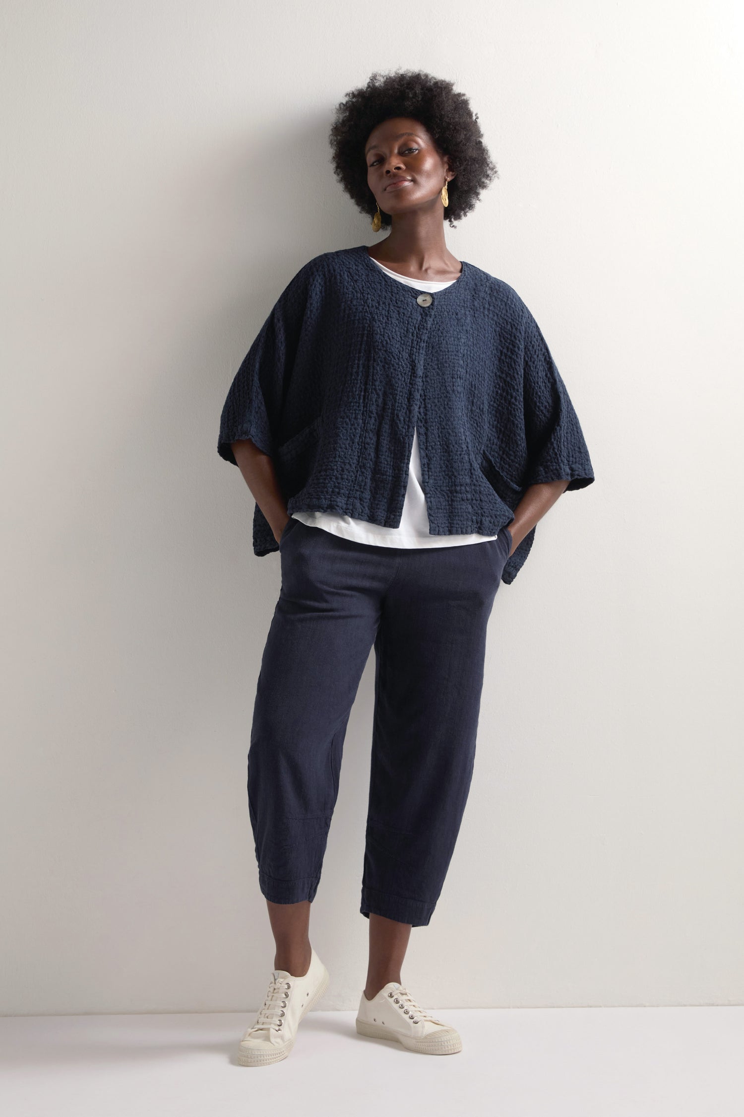 A person in a navy textured jacket and white shirt pairs Twisted Linen Crop Bubble Trousers with an elasticated waistband against a plain light background, finishing the look with crisp white sneakers.