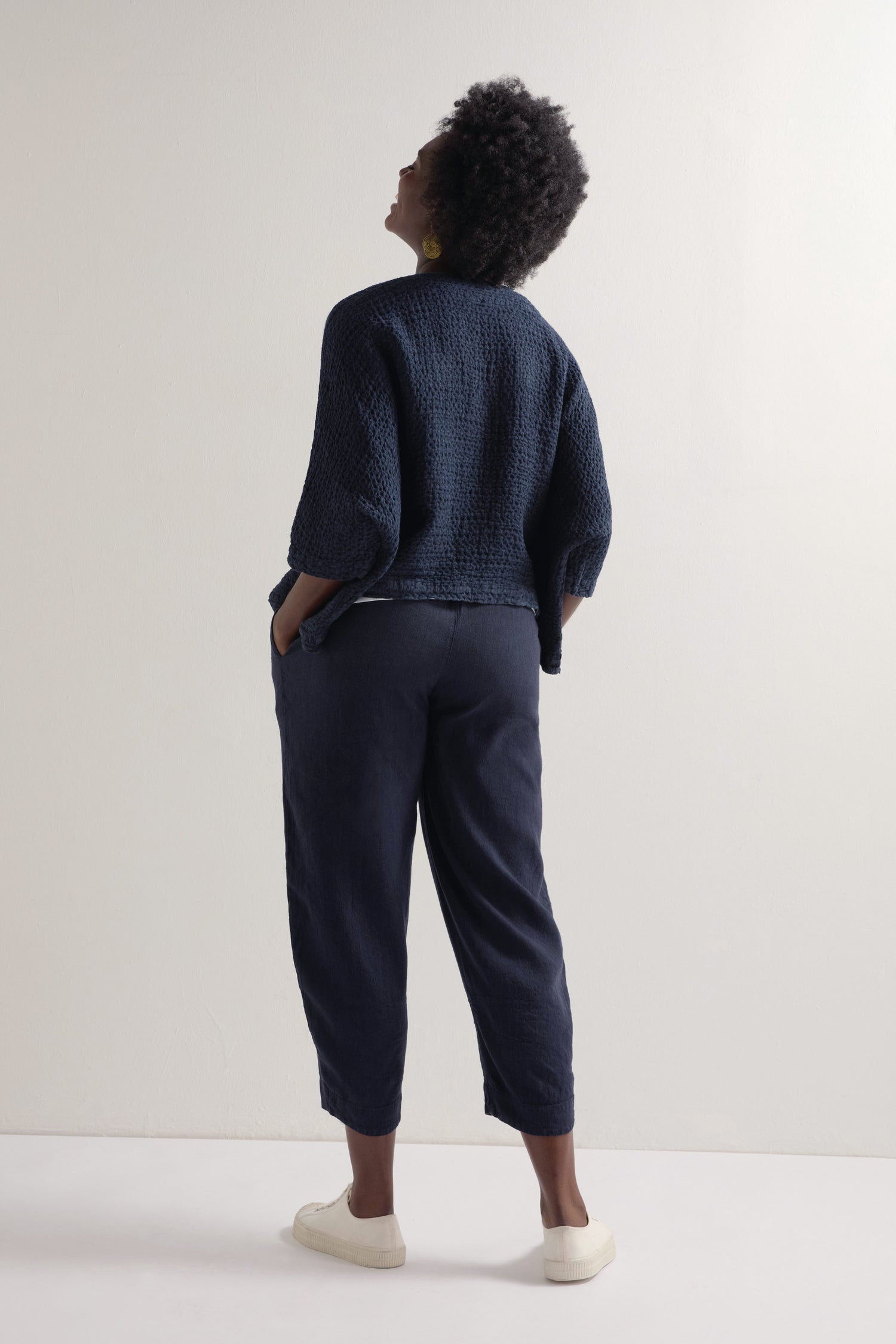 A person with curly hair, in a textured navy top and Twisted Linen Crop Bubble Trouser, featuring an elasticated waistband, pairs the outfit with white sneakers. They stand facing a light-colored wall, hands in pockets.