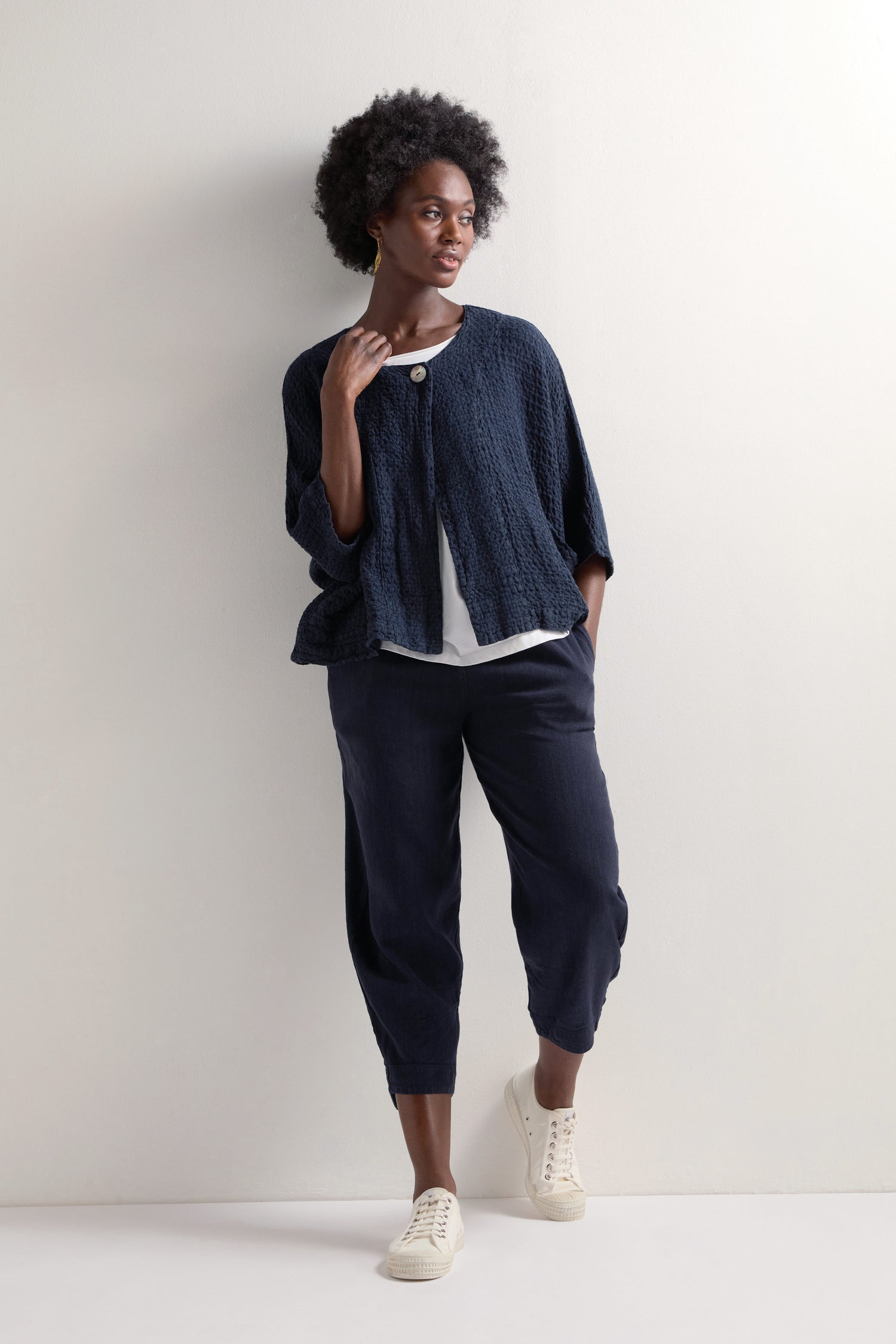 A person stands against a white wall wearing a navy blue textured cardigan, a white top, and Twisted Linen Crop Bubble Trousers with an elastic waistband, paired with white sneakers.