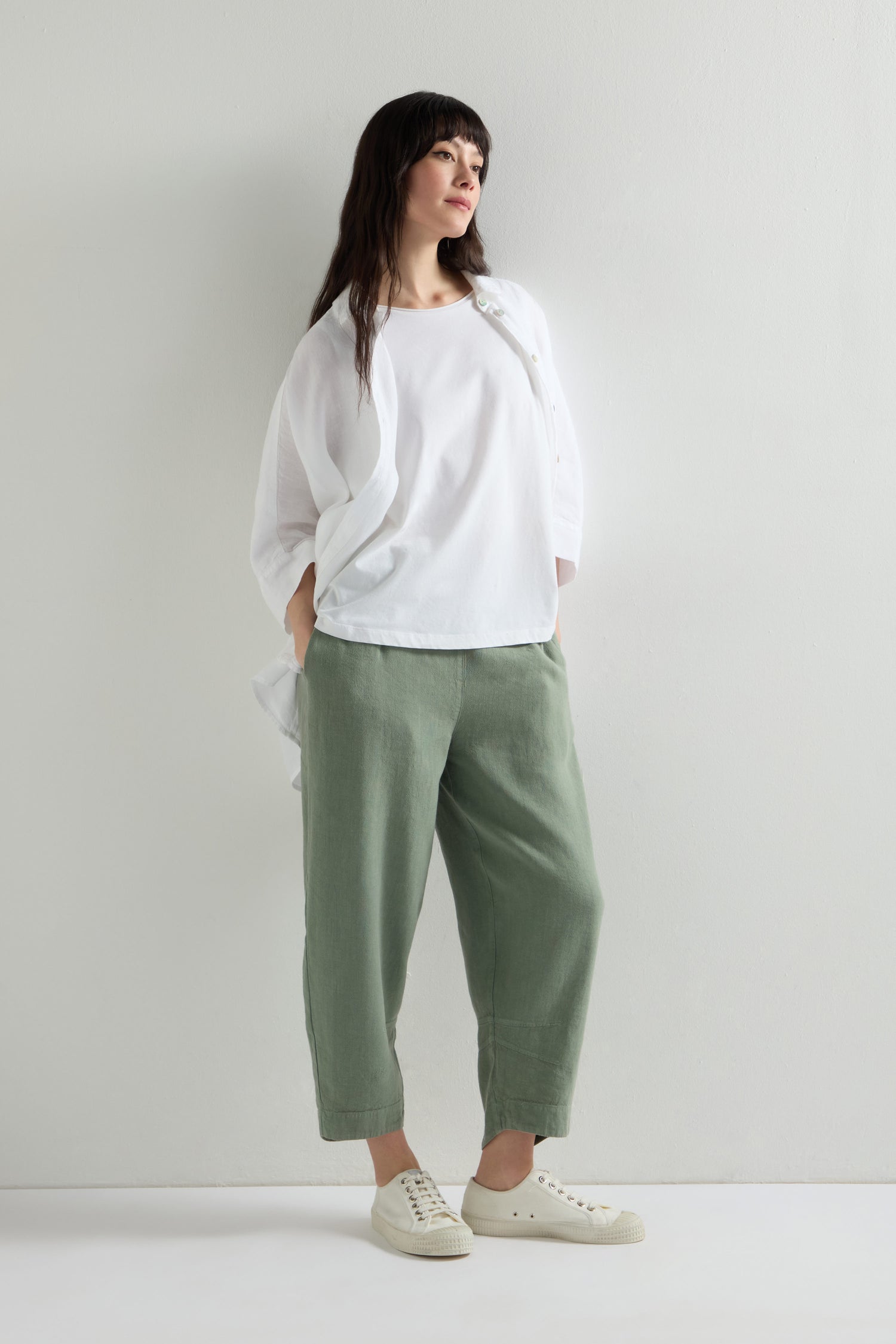 A woman in a white shirt, Twisted Linen Crop Bubble Trouser with an elastic waistband, and white sneakers stands with her hands in her pockets against a plain background.