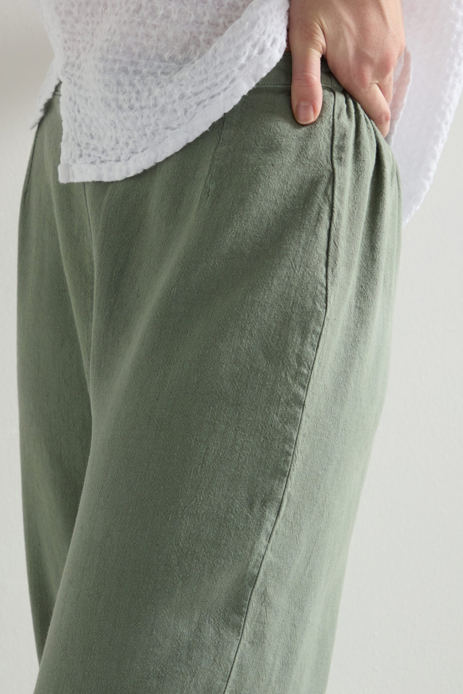 Close-up of a person wearing Twisted Linen Crop Bubble Trousers in light green, showcasing an elasticated waistband, a textured white top, and a hand tucked into the side pocket.