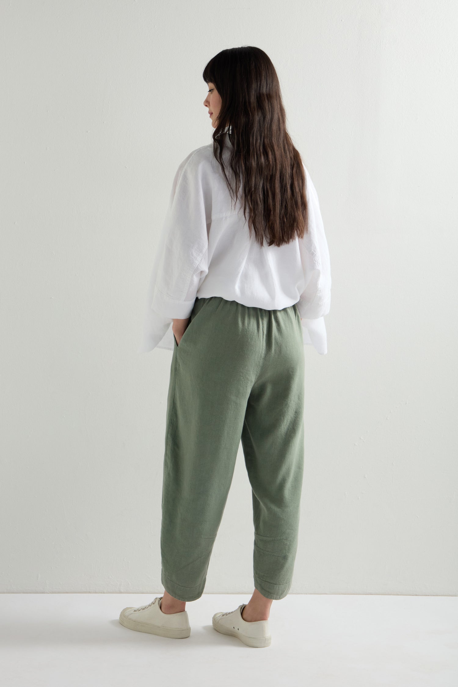 A person with long dark hair is wearing a white shirt, Twisted Linen Crop Bubble Trouser with an elasticated waistband, and white shoes, standing against a plain light background.