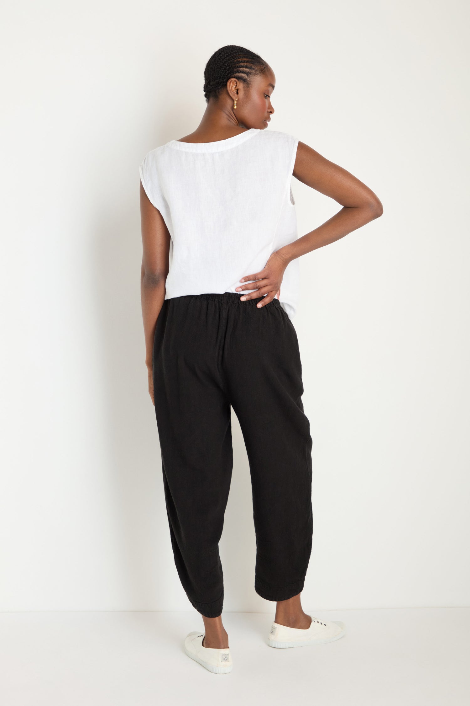 A person stands facing away, wearing a sleeveless white top, the Twisted Linen Crop Bubble Trouser in black, and white sneakers against a plain white background.