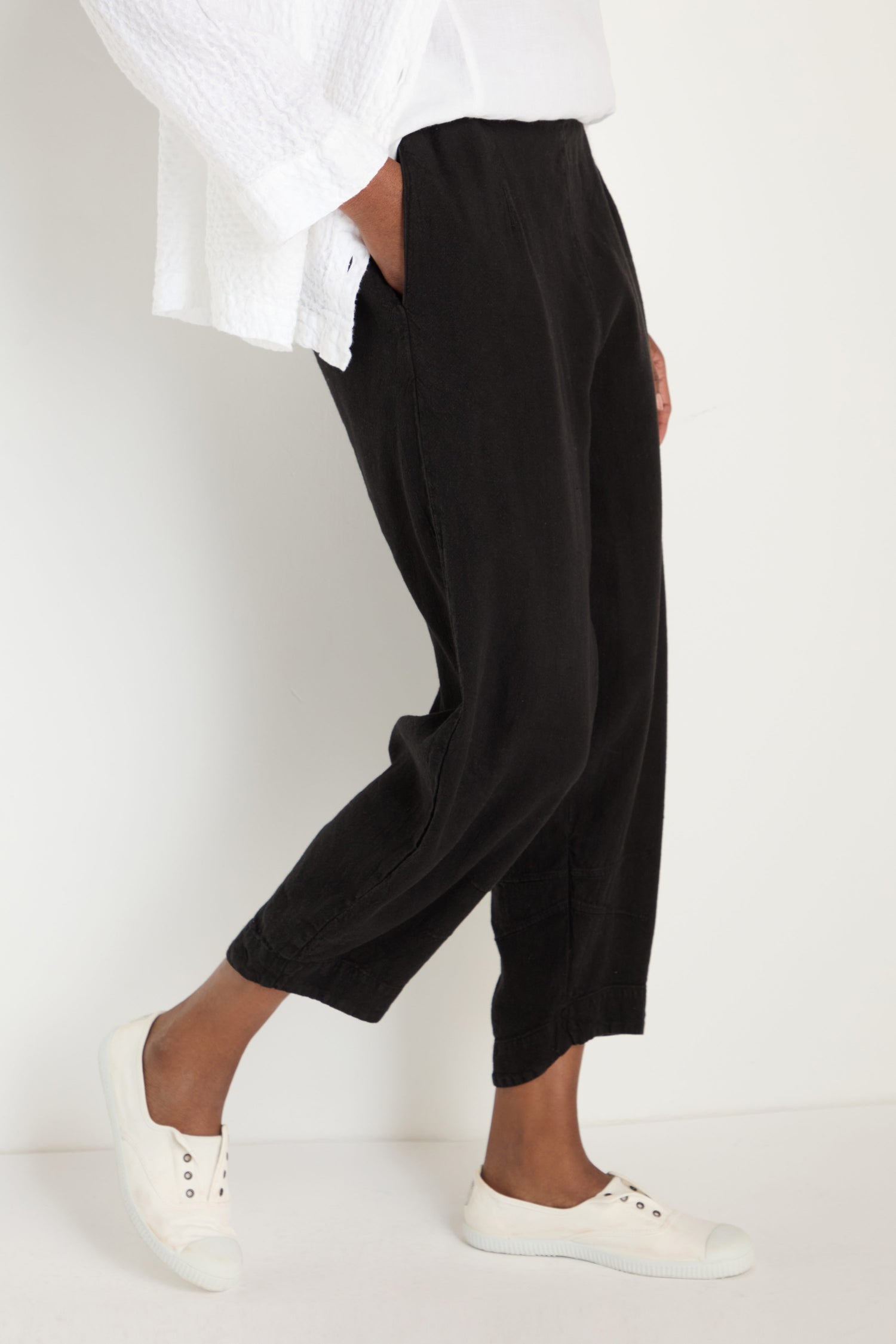 A person wearing black Twisted Linen Crop Bubble Trousers, a white textured top, and white slip-on shoes stands against a plain white background.