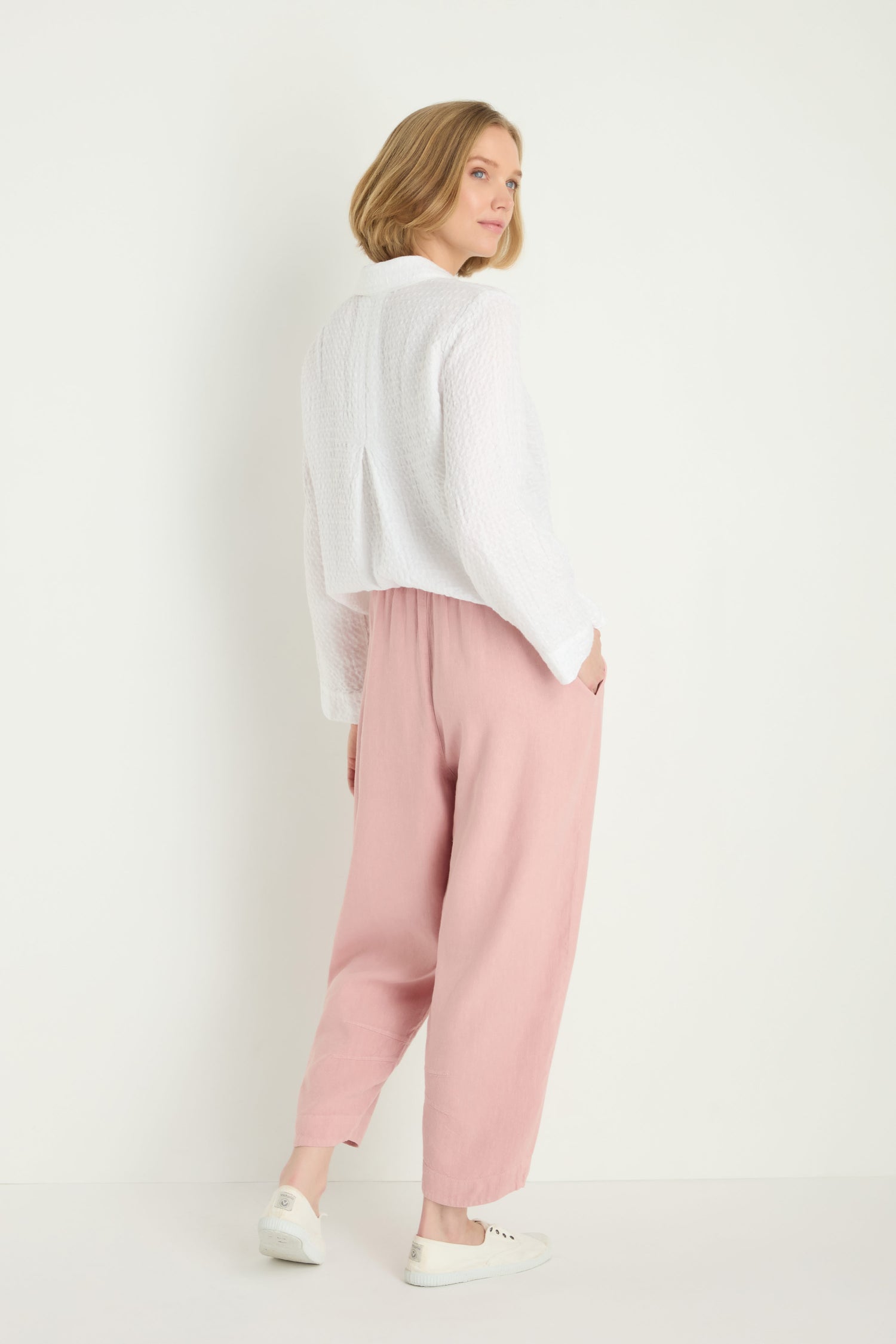 A woman stands slightly turned away, wearing a white textured long-sleeve top, the Twisted Linen Crop Bubble Trouser in pink for breathability, and white sneakers against a plain white background.