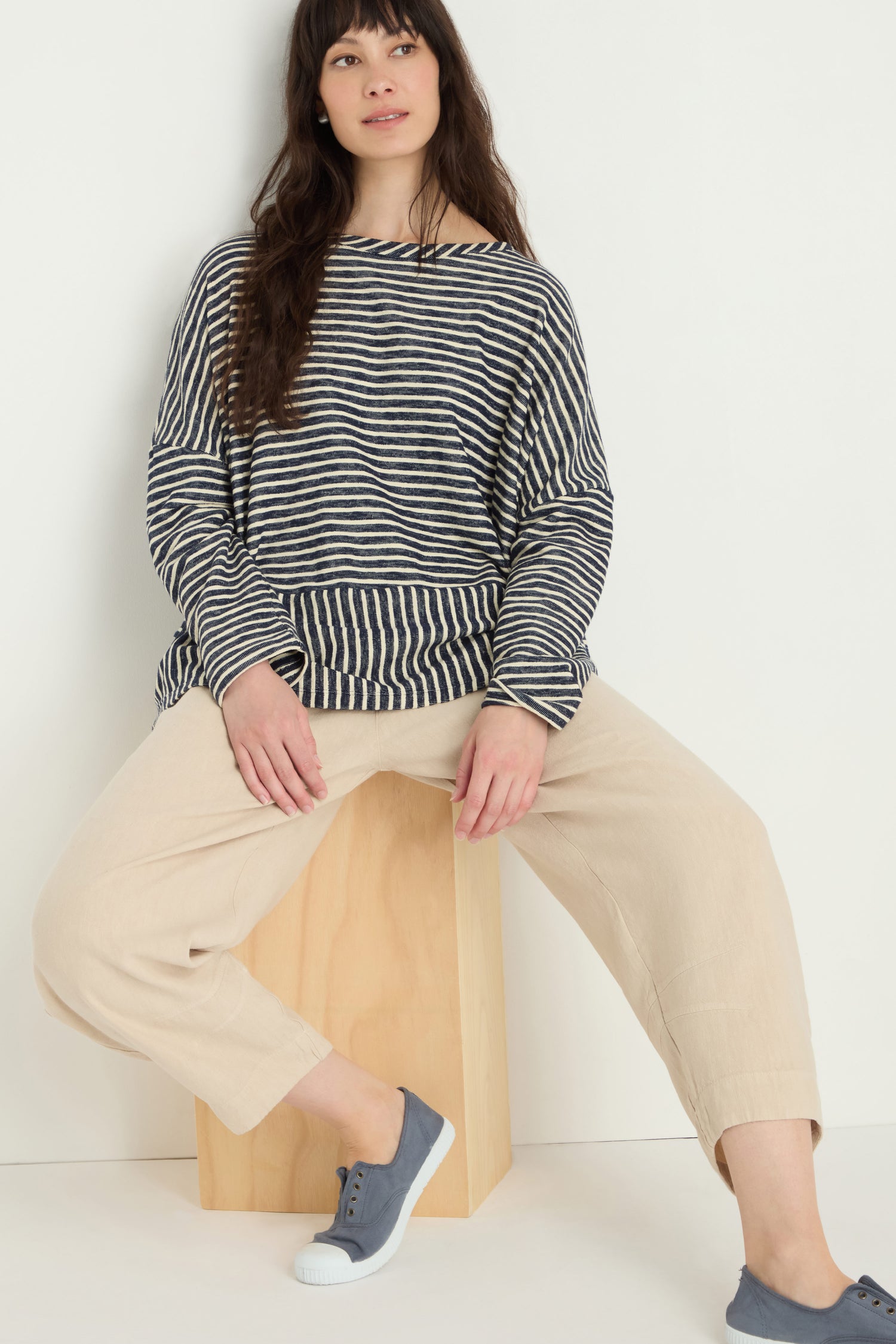 A woman wearing a black and white striped long-sleeve top, Twisted Linen Crop Bubble Trouser in beige, and blue sneakers sits on a wooden block against a plain white background.