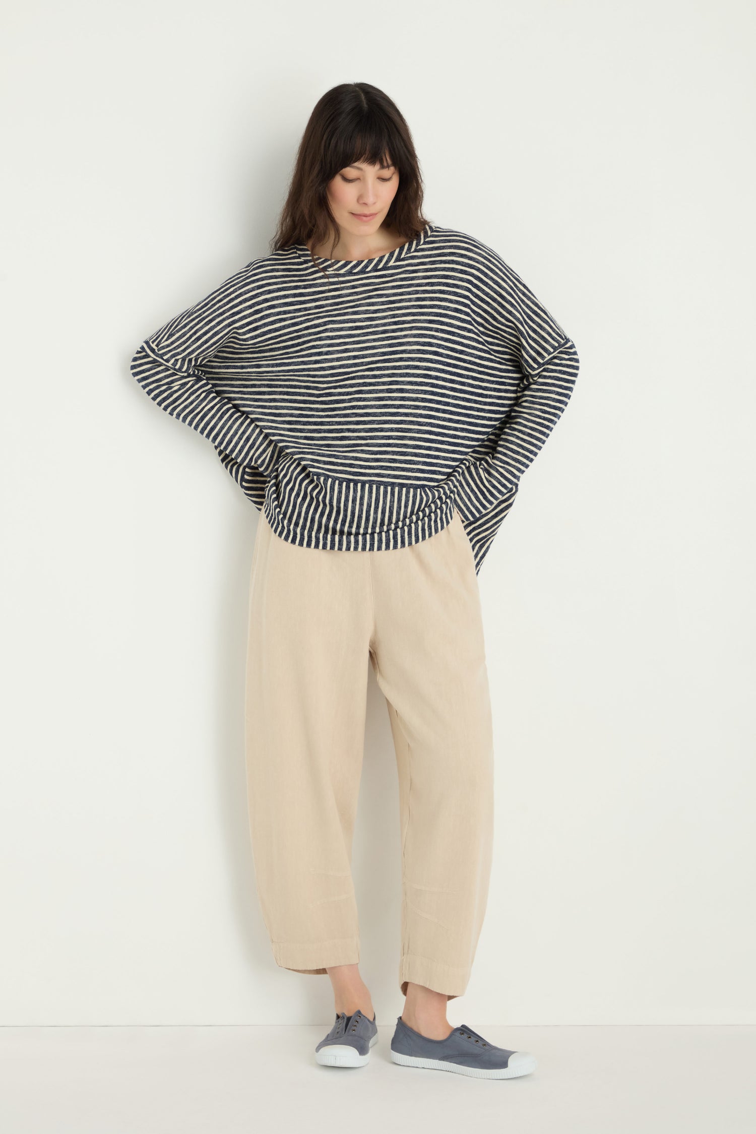 A person stands against a white background wearing a black and white striped long-sleeve top, Twisted Linen Crop Bubble Trouser in beige with a breathable feel, and gray sneakers, looking down with hands on hips.