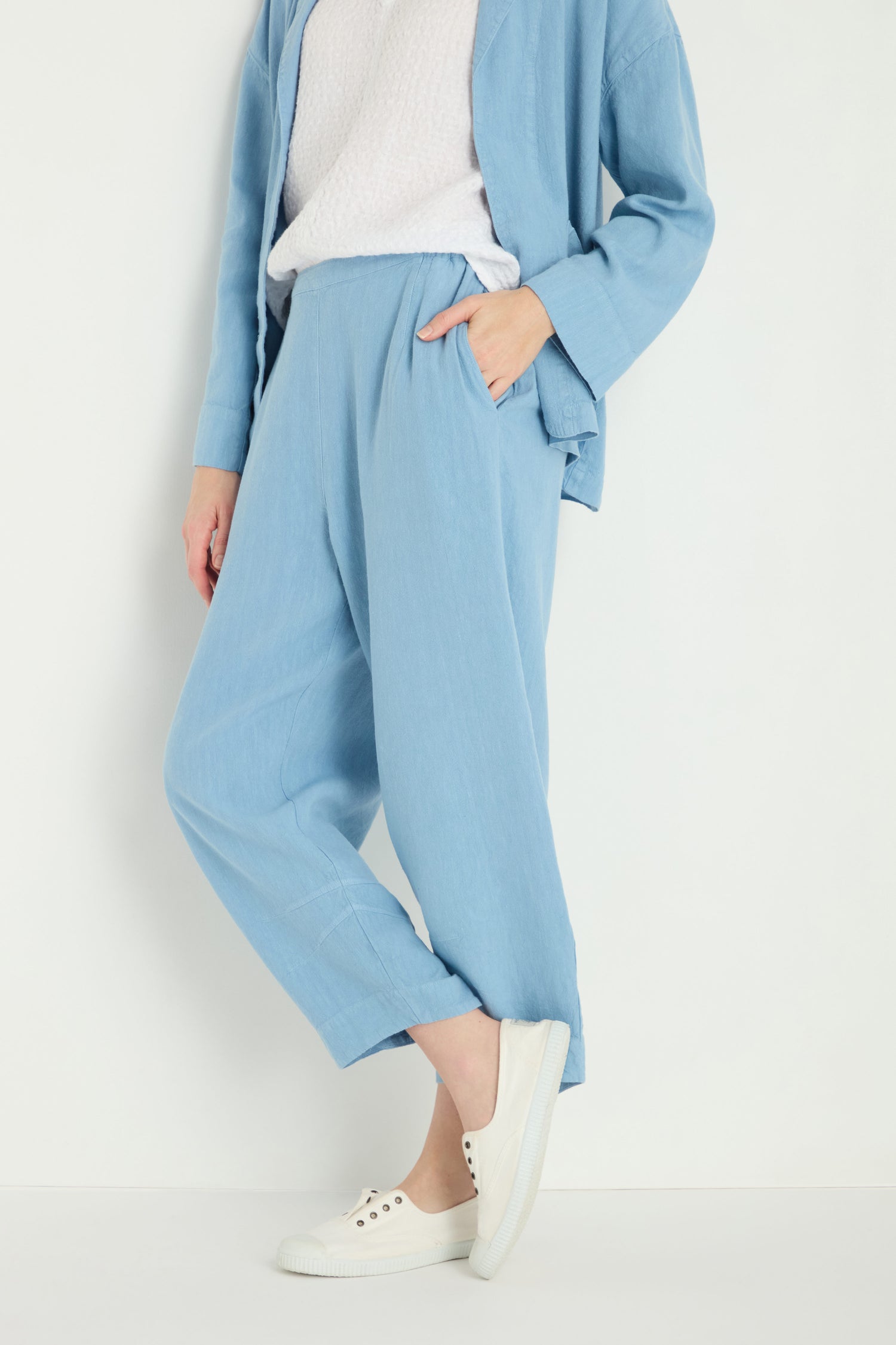 Wearing Twisted Linen Crop Bubble Trouser in light blue, paired with a matching jacket, white textured top, and white slip-on sneakers, standing against a plain light background.