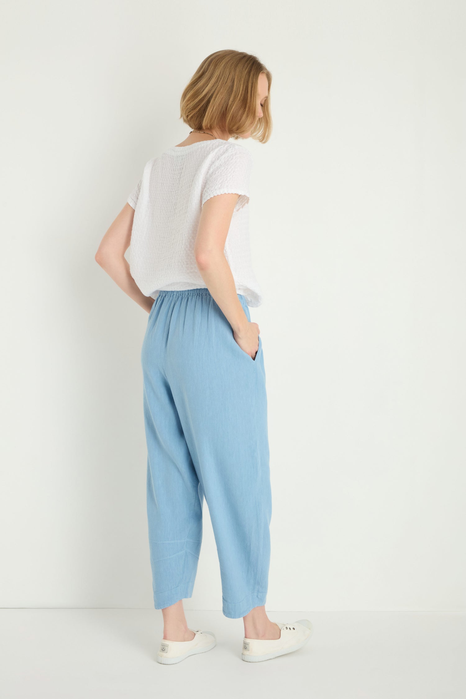 A person stands facing away, wearing a white short-sleeve top, Twisted Linen Crop Bubble Trousers in light blue, and white sneakers against a plain white background.