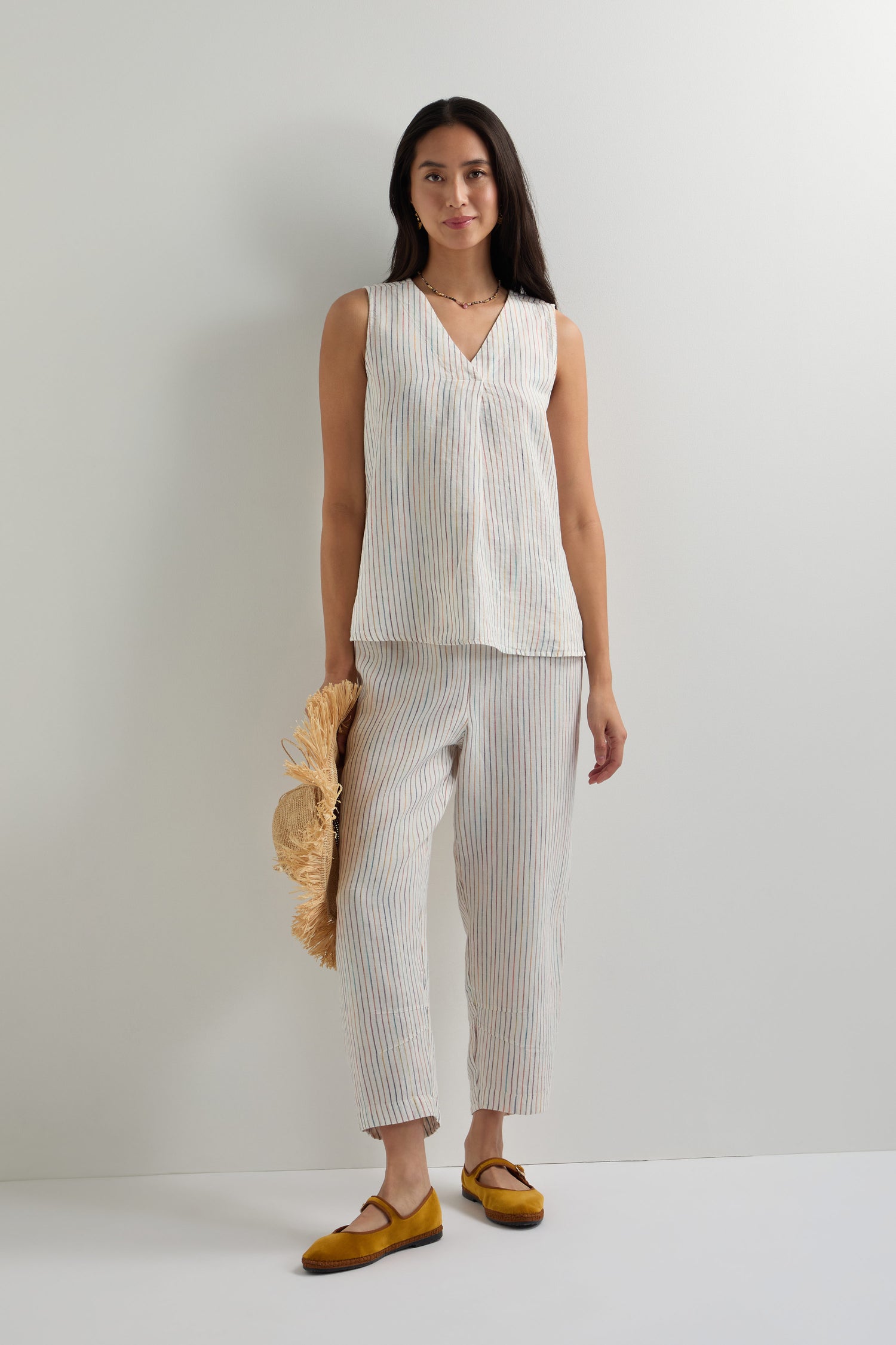Woman stands against a plain wall wearing a sleeveless striped top and White Space Dye Stripe Bubble Trousers, holding a straw hat, and wearing mustard-colored flats.