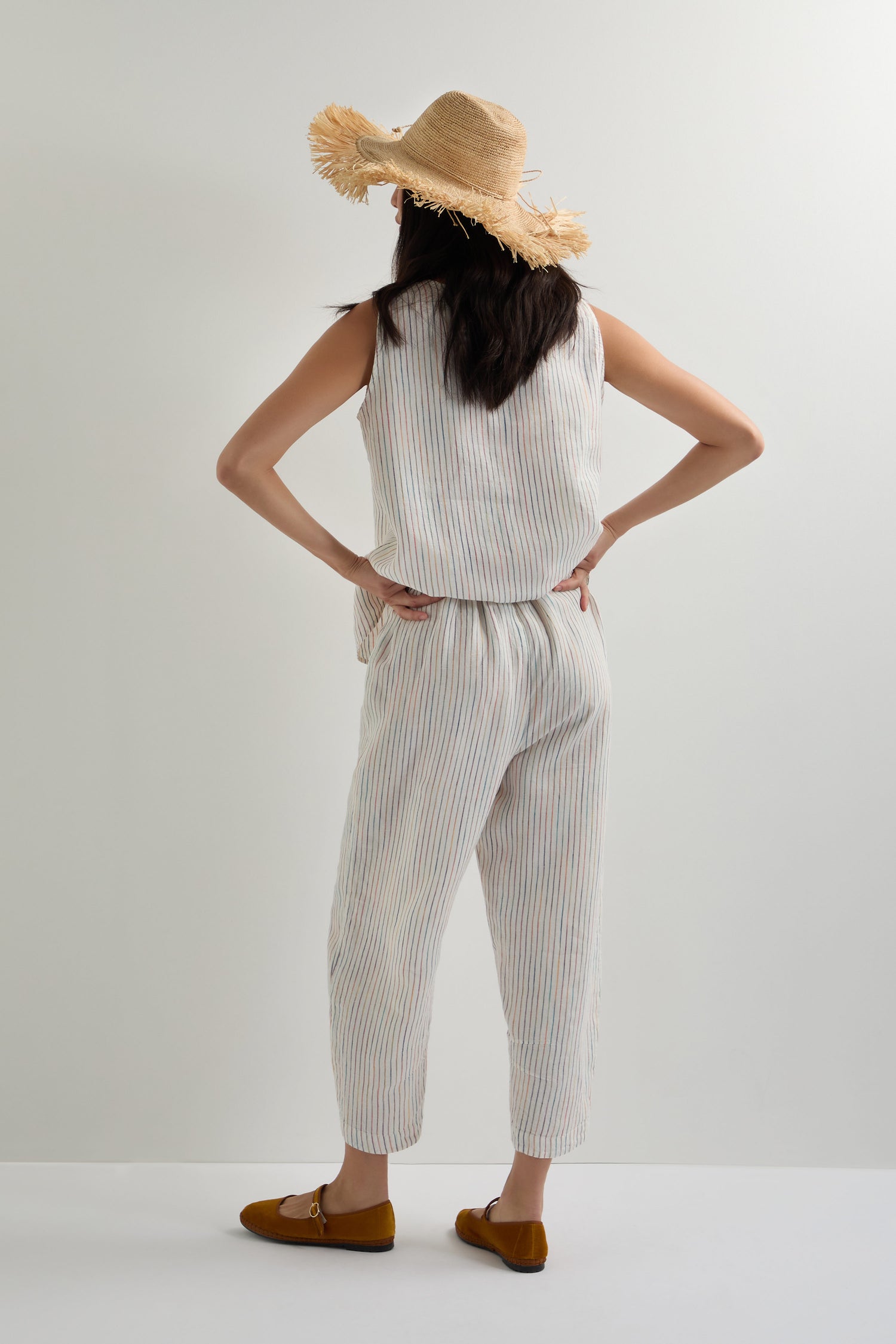 A person stands with hands on hips, facing away from the camera, wearing a straw hat and the White Space Dye Stripe Bubble Trouser with a bubble silhouette, sleeveless top, and brown flat shoes against a plain background.