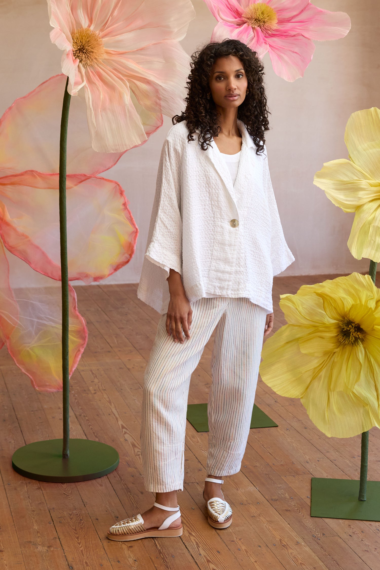 A woman in a textured white jacket and White Space Dye Linen Stripe Bubble Trouser stands indoors between large decorative flower sculptures on a wooden floor.