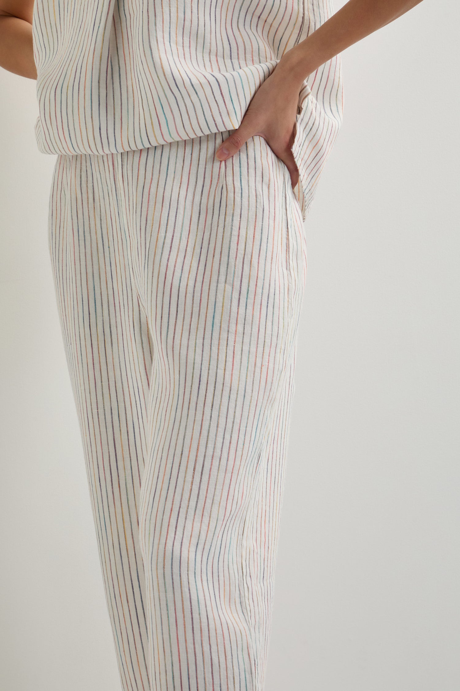 A person stands with one hand in their pocket, wearing the White Space Dye Stripe Bubble Trouser—a matching set of white clothes featuring thin, multicolored vertical stripes and cropped linen trousers.