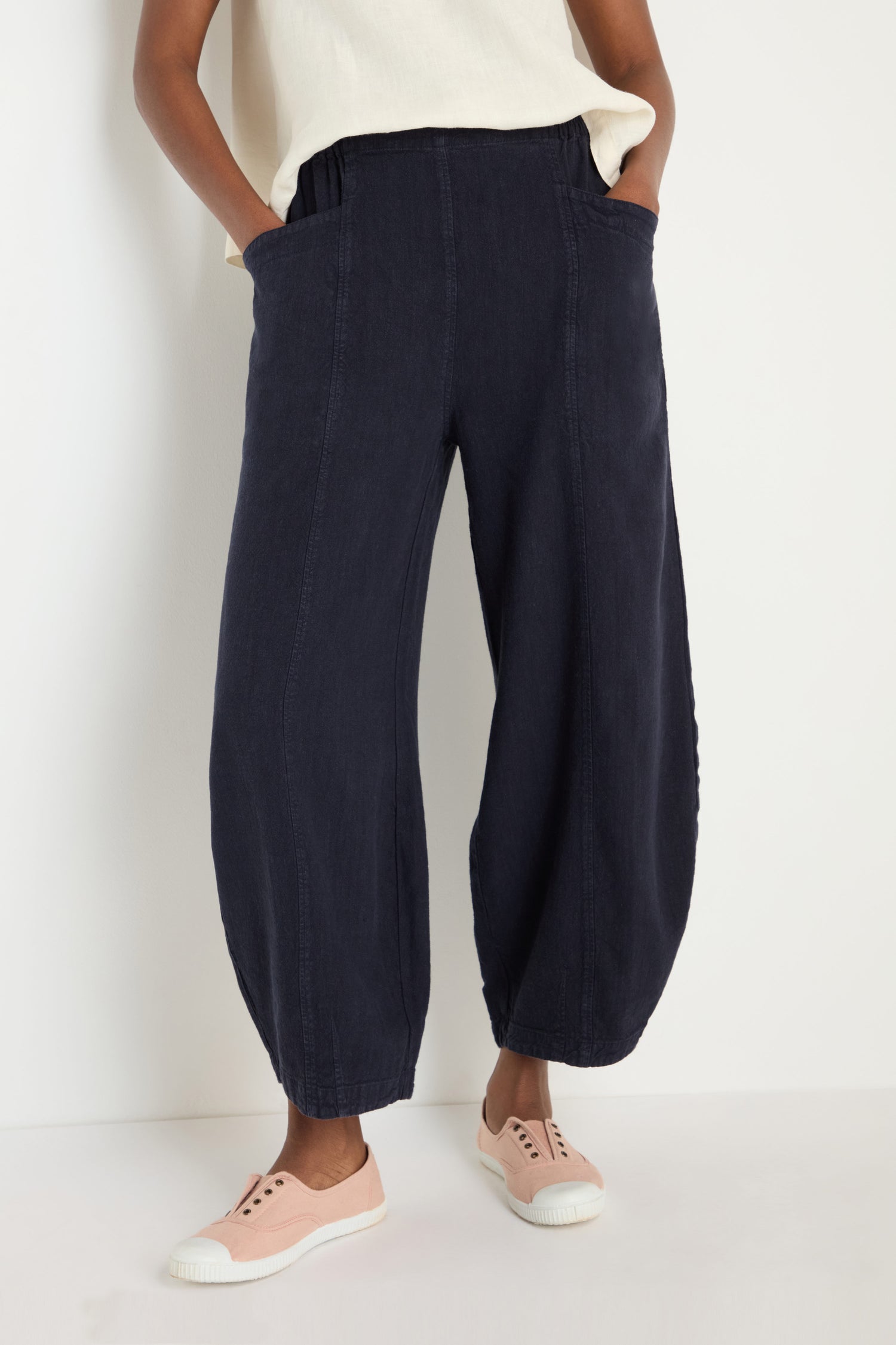 A person stands against a white background, hands in pockets, wearing the Twisted Linen Bubble Trouser in navy blue with a white top and pale pink sneakers. The trouser’s airy linen fabric offers both comfort and style.
