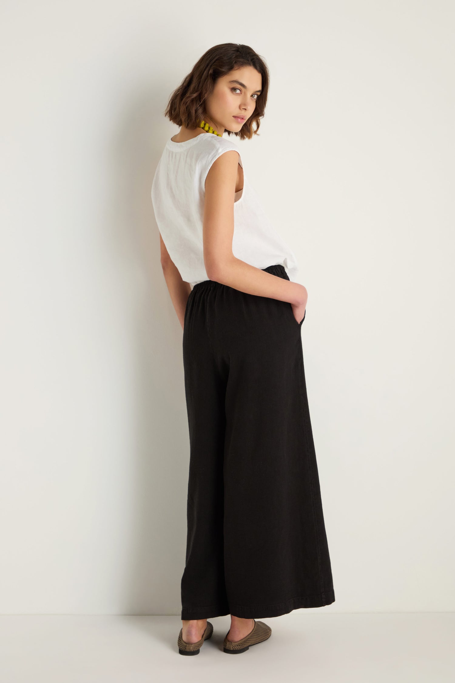 A woman stands against a plain light wall, wearing a sleeveless white top, the Twisted Linen Wide Trouser in black, and brown shoes, with one hand in her pocket and looking over her shoulder.