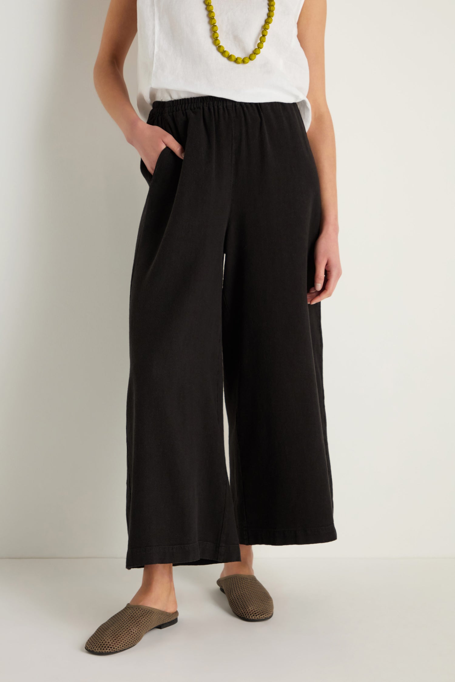 A person stands against a plain light background wearing a sleeveless white top, yellow bead necklace, brown slip-on shoes, and the Twisted Linen Wide Trouser featuring a textured linen blend and pockets.