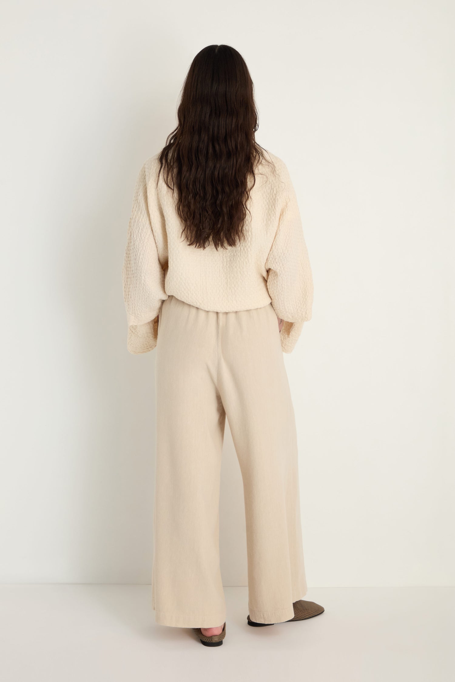 A person with long brown hair stands facing away, wearing a cream textured sweater and Twisted Linen Wide Trouser, against a plain white background.