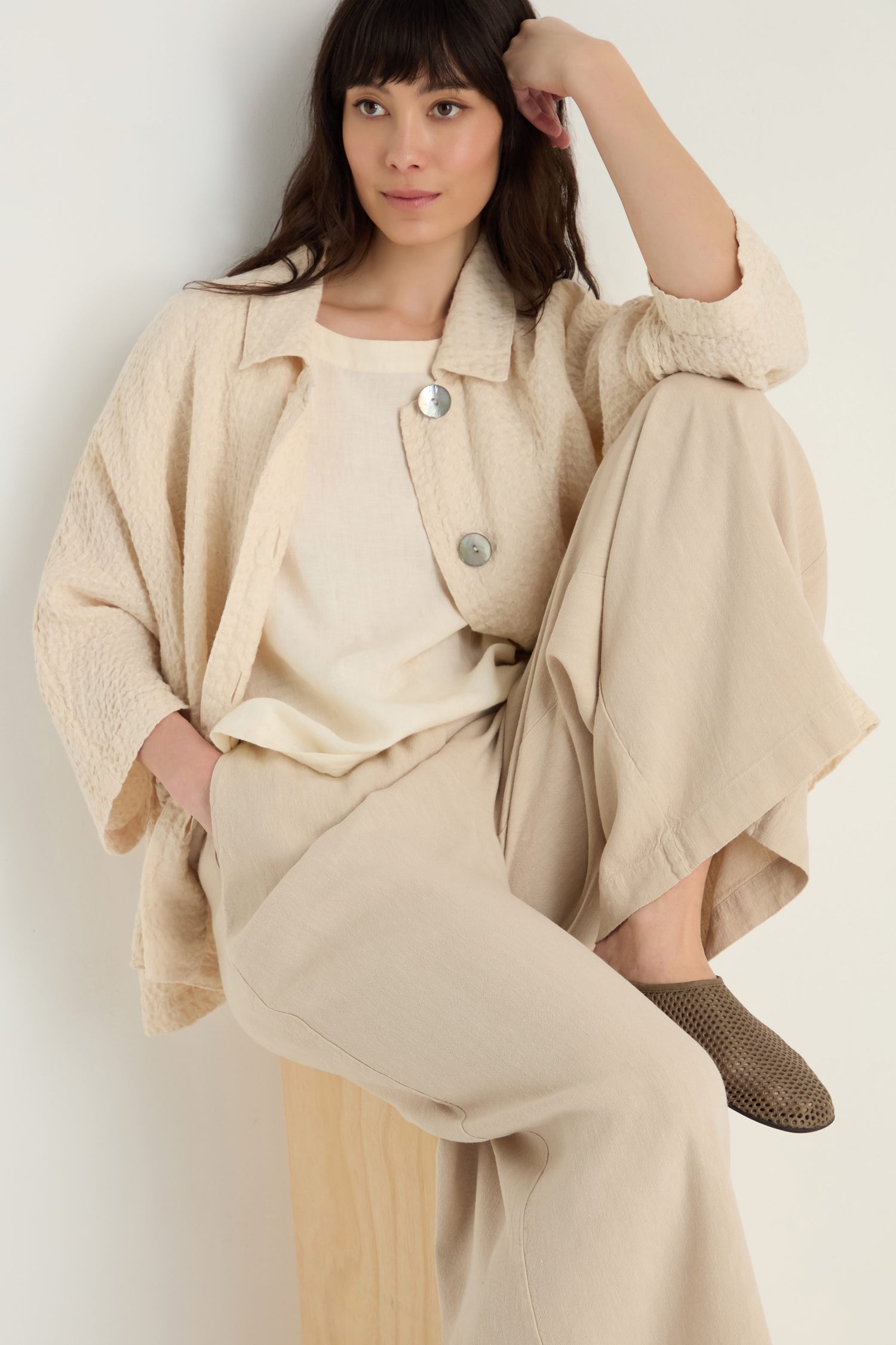 A woman sits on a wooden block, wearing a cream-textured jacket, Twisted Linen Wide Trouser, brown woven shoes, and rests her left arm on her raised knee.