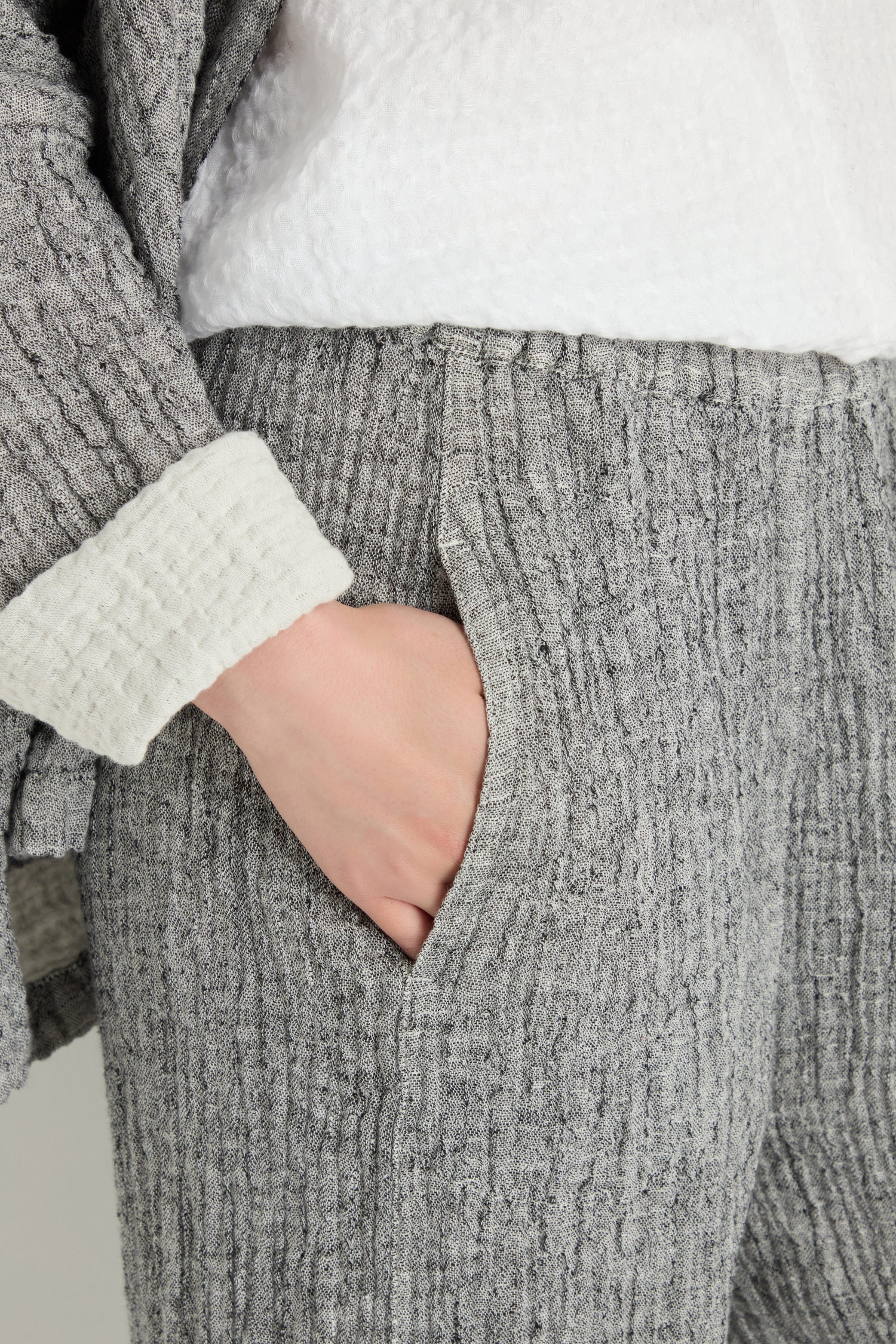 A person wears the Crinkle Double Face Linen Bubble Trouser in textured grey linen with a white top and has their hand in the pocket.