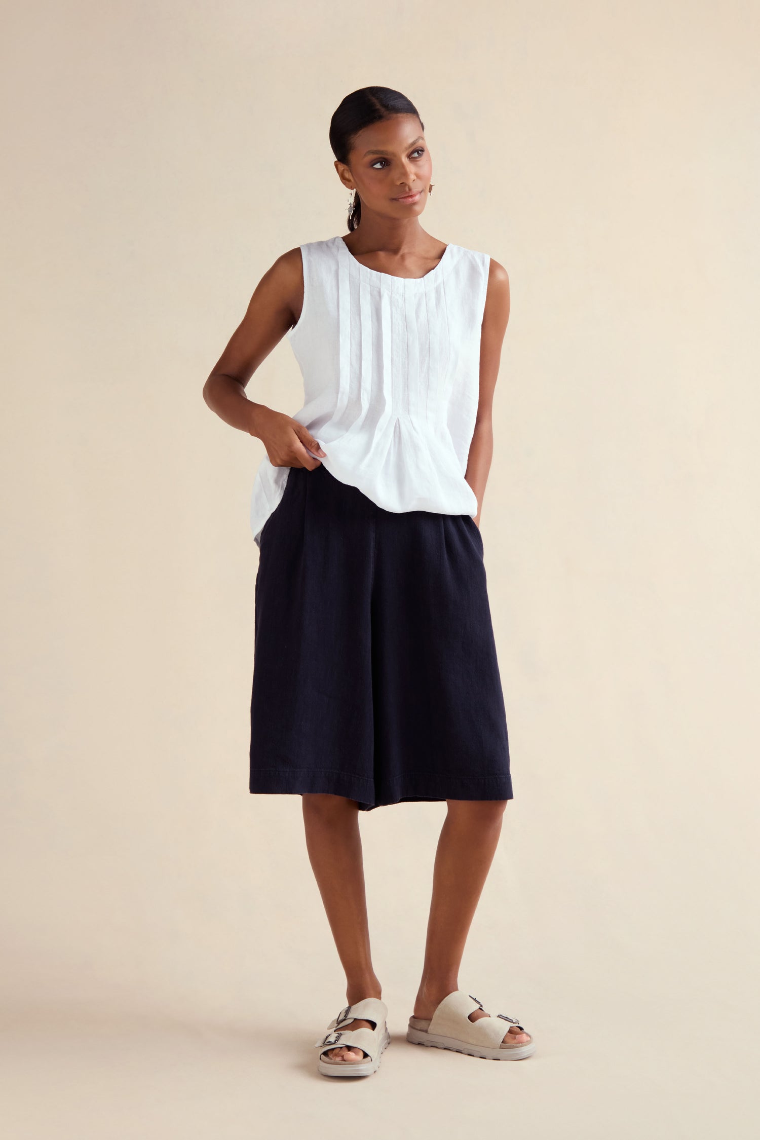A woman stands against a plain background wearing a sleeveless white top, Twisted Linen Wide Shorts in black, and light-colored sandals.