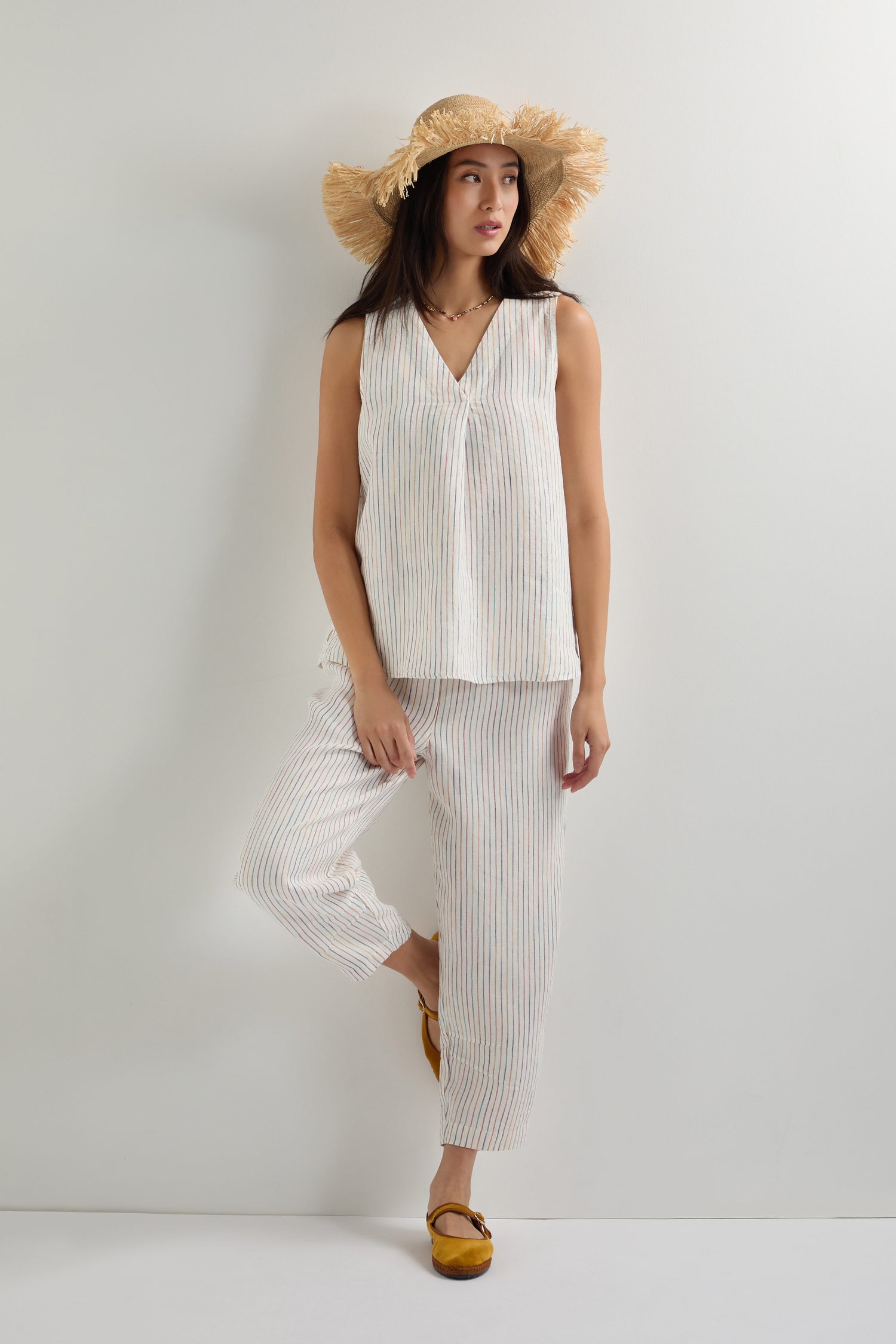 A woman in a straw hat and an oversized White Space Dye Stripe Linen Vest with matching loose pants stands against a plain light wall, her feet crossed.