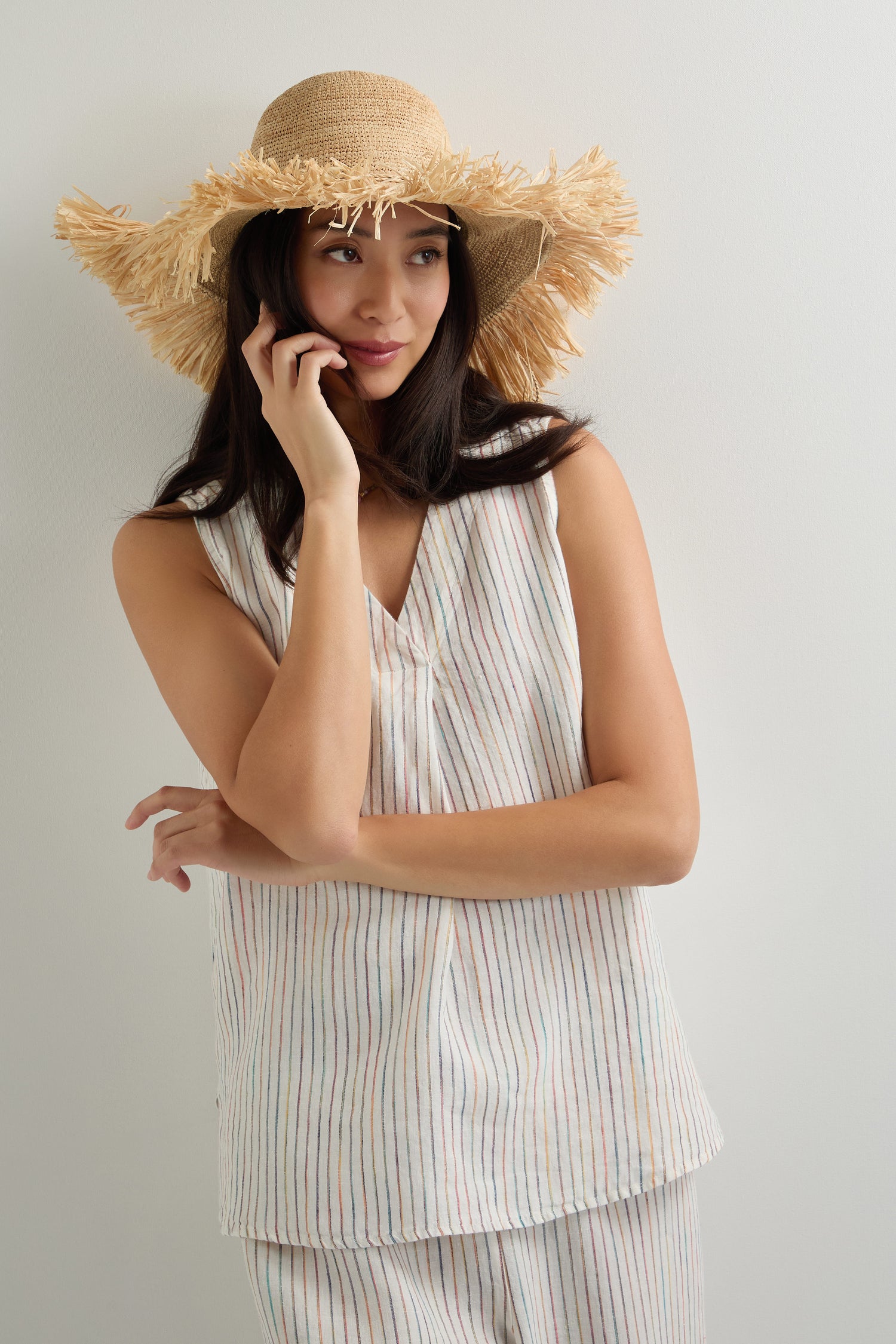 A woman wearing a wide-brimmed straw hat and an oversized White Space Dye Stripe Linen Vest stands against a plain wall, gazing to the side with one hand on her face.