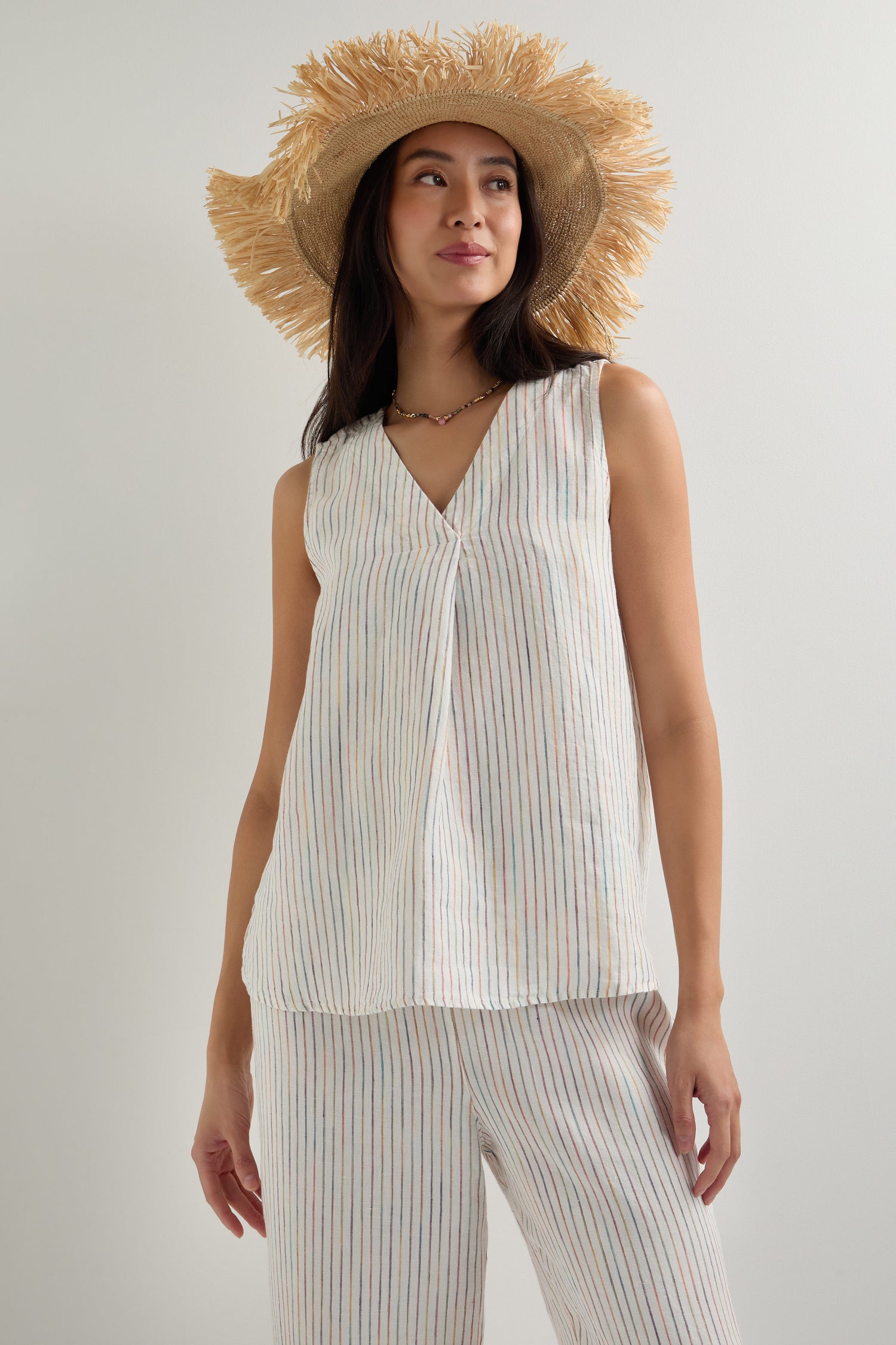 A woman in a straw hat and an oversized White Space Dye Stripe Linen Vest with matching striped pants stands against a plain light background.